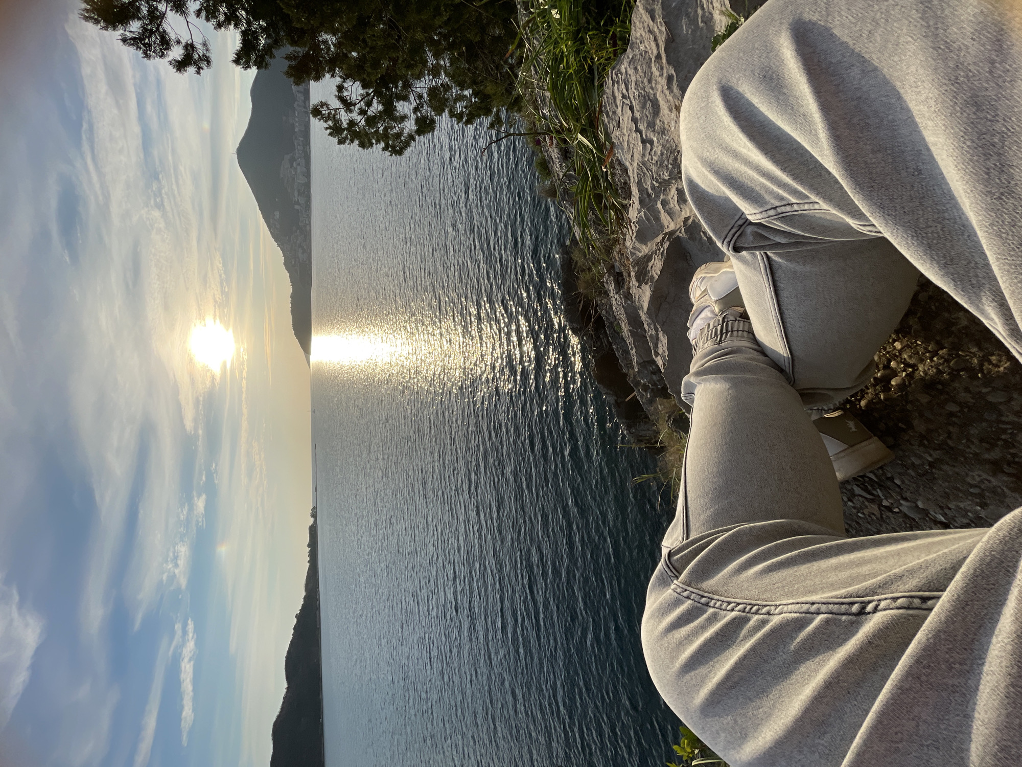 The image captures a first-person perspective from an elevated rocky outcrop overlooking a large body of water, identified as the Adriatic Sea in the Budva Bay area of Montenegro, likely a viewpoint west of Budva Old Town towards Mogren Beach.
In the immediate foreground, the lower half of a person's legs is visible, clad in light grey, possibly acid-wash, denim jeans and light-colored athletic shoes with dark accents. The legs are crossed at the ankles, indicating a seated or reclining position. The ground beneath the legs is composed of textured grey rock and scattered small pebbles, interspersed with sparse green vegetation, including grass and low-lying plants. Dark green coniferous foliage partially frames the right side of the scene.
Beyond the immediate foreground, a calm, dark blue expanse of water stretches towards the horizon. The sun, appearing as a bright white-yellow orb, is low in the sky, positioned in the upper right-center, casting a distinct shimmering golden reflection path across the water's surface towards the viewer. The sky above is a clear light blue with scattered white, wispy clouds. A faint, arc-shaped optical phenomenon, consistent with a sun dog or halo, is discernible to the left of the sun.
Geographically, a dark, lower-lying landmass extends into the water on the far left. To the right, a prominent, higher mountain, identified as Mount Spas, rises from the coastline. Its lower slopes and base are visibly covered with numerous white and light-colored buildings, forming the urban area of Budva. A distinct antenna or communication tower is visible near the summit of Mount Spas. On the far-left horizon, a small white vessel, consistent with a sailboat, is faintly visible on the water.
