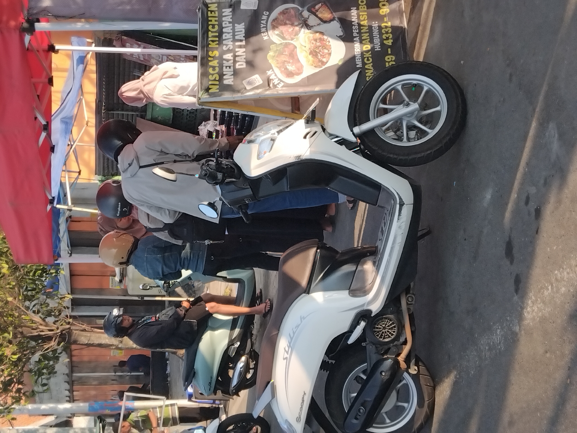 This image depicts a street scene in Karanganyar, Indonesia, under a bright, likely morning or late afternoon sun, casting long shadows. In the foreground, a white Honda Scoopy motorcycle is parked, its "Stylish" branding visible on the seat. The motorcycle has a black exhaust and a silver wheel. To the right of the motorcycle, an A-frame sign stands, advertising "NISCA'S KITCHEN ANEKA SARAPAN DAN LAUK" (Nisca's Kitchen Various Breakfasts and Side Dishes). The sign also features images of food, the text "ENAK" (delicious) and "KENYANG" (full), and contact information including a phone number. Behind the Scoopy, a light blue scooter is parked, with a person wearing black shorts and flip-flops sitting on it, their legs visible. A group of people are gathered near a red canopy tent. One individual, wearing a black helmet and a grey hooded jacket, is looking down at something. Another person in a denim jacket and a tan helmet is also present. A woman wearing a light pink headscarf is standing near the sign. The background shows a building with corrugated metal siding and a tree with sparse leaves. The overall environment appears to be an outdoor market or roadside stall area.