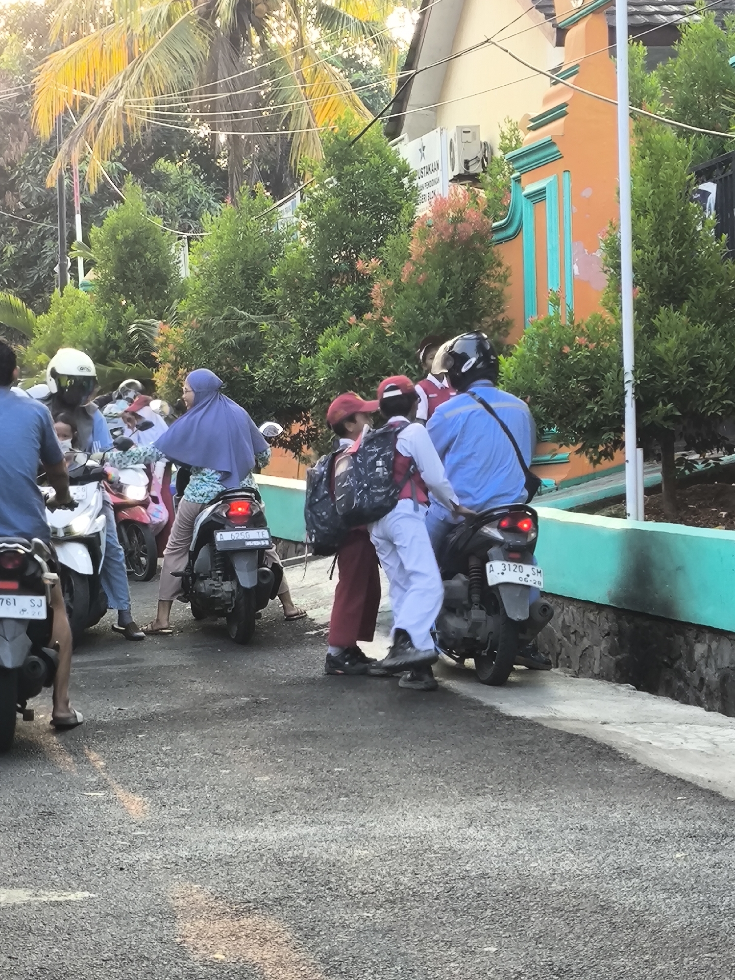 The image depicts a street scene in Cilegon, Indonesia, likely near a school given the presence of students in uniform. Several people are on or near motorcycles. In the foreground, a person wearing a blue shirt and jeans rides a motorcycle, facing away from the camera. To their right, a person wearing a red cap and white pants, carrying a backpack, stands next to another motorcycle. Behind them, a person in a blue shirt and jeans also rides a motorcycle, carrying a passenger in a purple headscarf and patterned shirt. Further in the background, a white motorcycle is parked, and other individuals are visible. The environment features trees, a building with orange and teal accents, and a stone wall. The road is asphalt, and sunlight casts shadows, suggesting it is daytime. The license plates visible on the motorcycles read "A 625G TE" and "A 3120 SM".