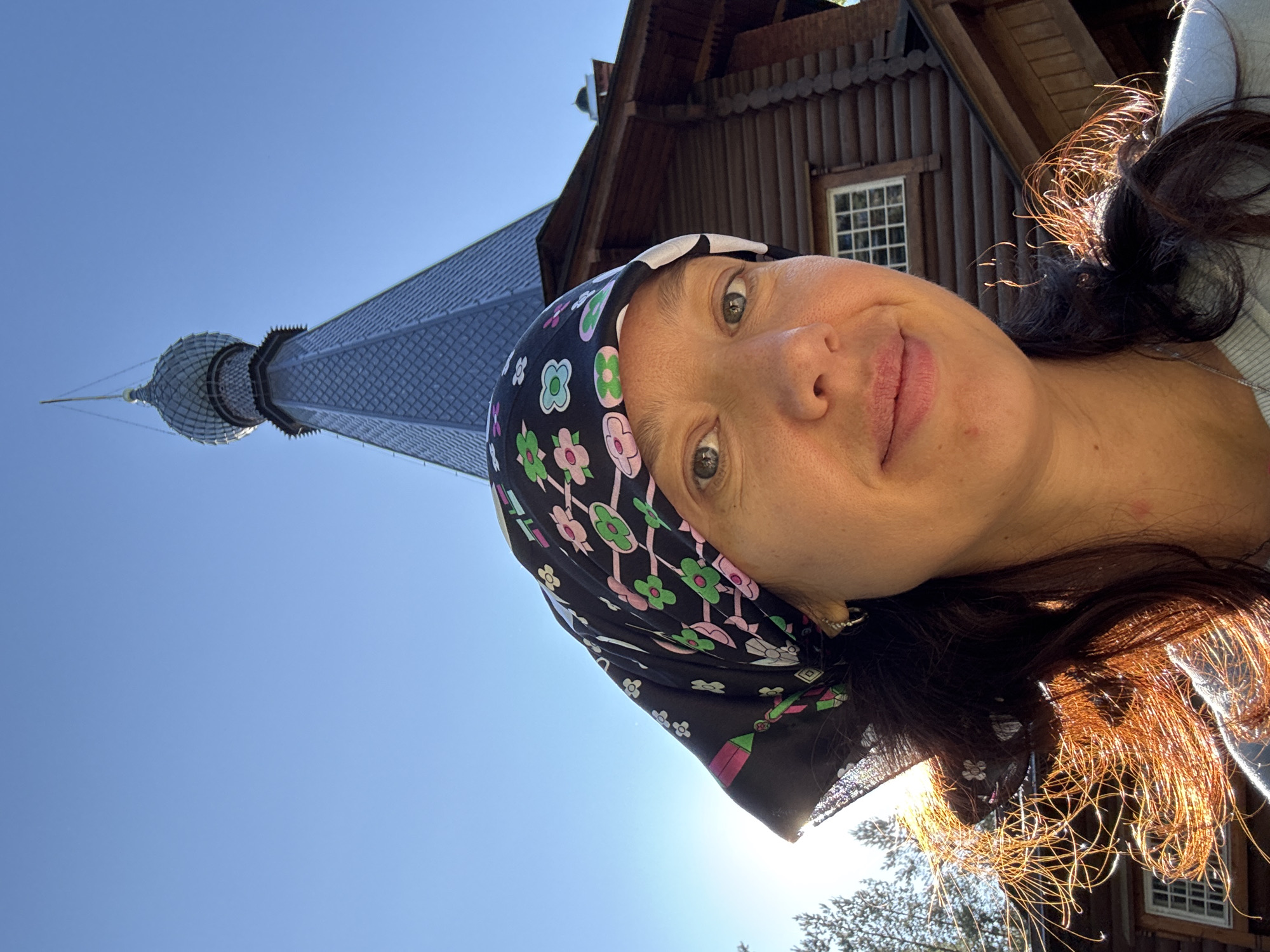 This is a portrait-style selfie taken outdoors, in front of a wooden building with a domed structure. The subject is a woman with dark hair, wearing a headscarf with a floral pattern and hoop earrings. She is looking towards the camera with a slight smile. Her hair is illuminated by the sun, creating a halo effect. Behind her, a large, dark wooden building with a distinctive grey, domed roof topped with a spire is visible. The building appears to be a church or chapel, consistent with the architecture found in Vyritsa, Russia, a town known for its numerous religious sites. The sky is clear and blue, indicating a sunny day. The overall scene suggests a visit to a historic or religious location in Vyritsa.