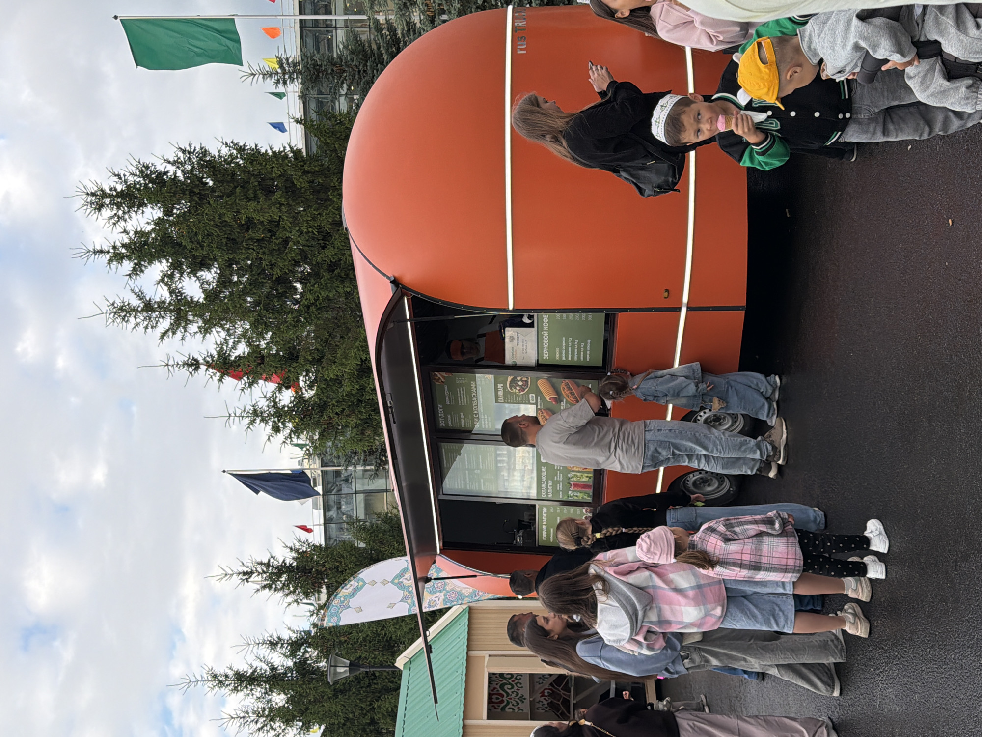 An outdoor scene depicts a food truck with an orange, rounded body parked on a wet asphalt surface. Several people, including adults and children, are gathered around the food truck, some in line and others interacting. The food truck has a large window displaying a menu, and a vendor is visible inside. Flags are seen in the background, along with evergreen trees and a building. One child is eating an ice cream cone, and another child is seated in what appears to be a stroller or carrier. The sky is overcast.