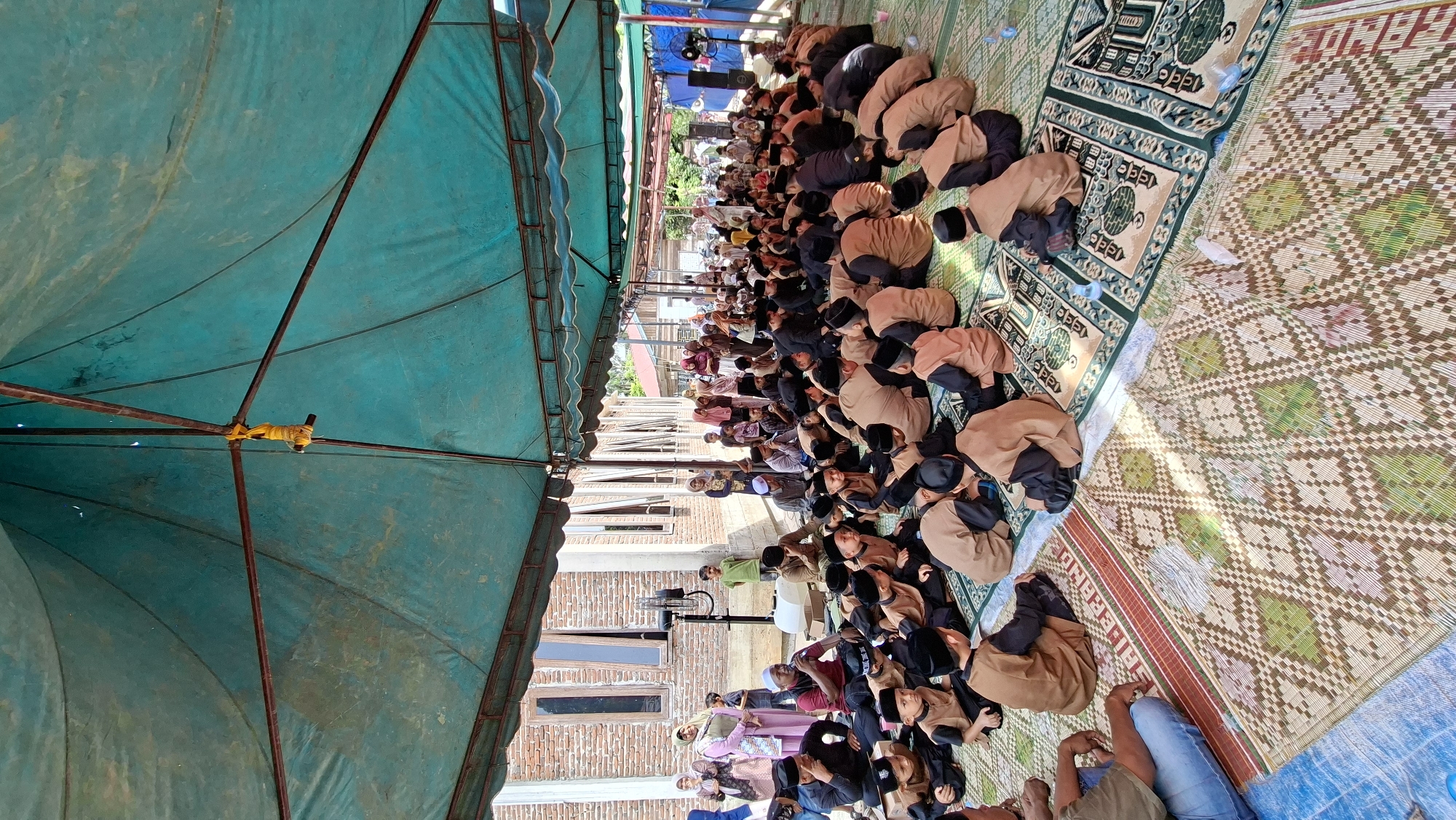 This image depicts an outdoor gathering under a large, teal-colored tent, likely for a community event or religious ceremony. The scene appears to be in Asan, Indonesia, indicated by the context provided. A significant number of people, predominantly children wearing traditional Islamic attire including black skullcaps and light brown robes, are seated on woven mats and prayer rugs. They are arranged in rows and appear to be listening or engaged in an activity. In the background, adults are also present, some standing and others seated. A brick building with visible windows is situated behind the gathering. The environment suggests a sunny day, with the tent providing shade. Objects visible include a portable fan, speakers, and water bottles scattered among the attendees. The overall atmosphere seems to be one of communal assembly and shared activity.