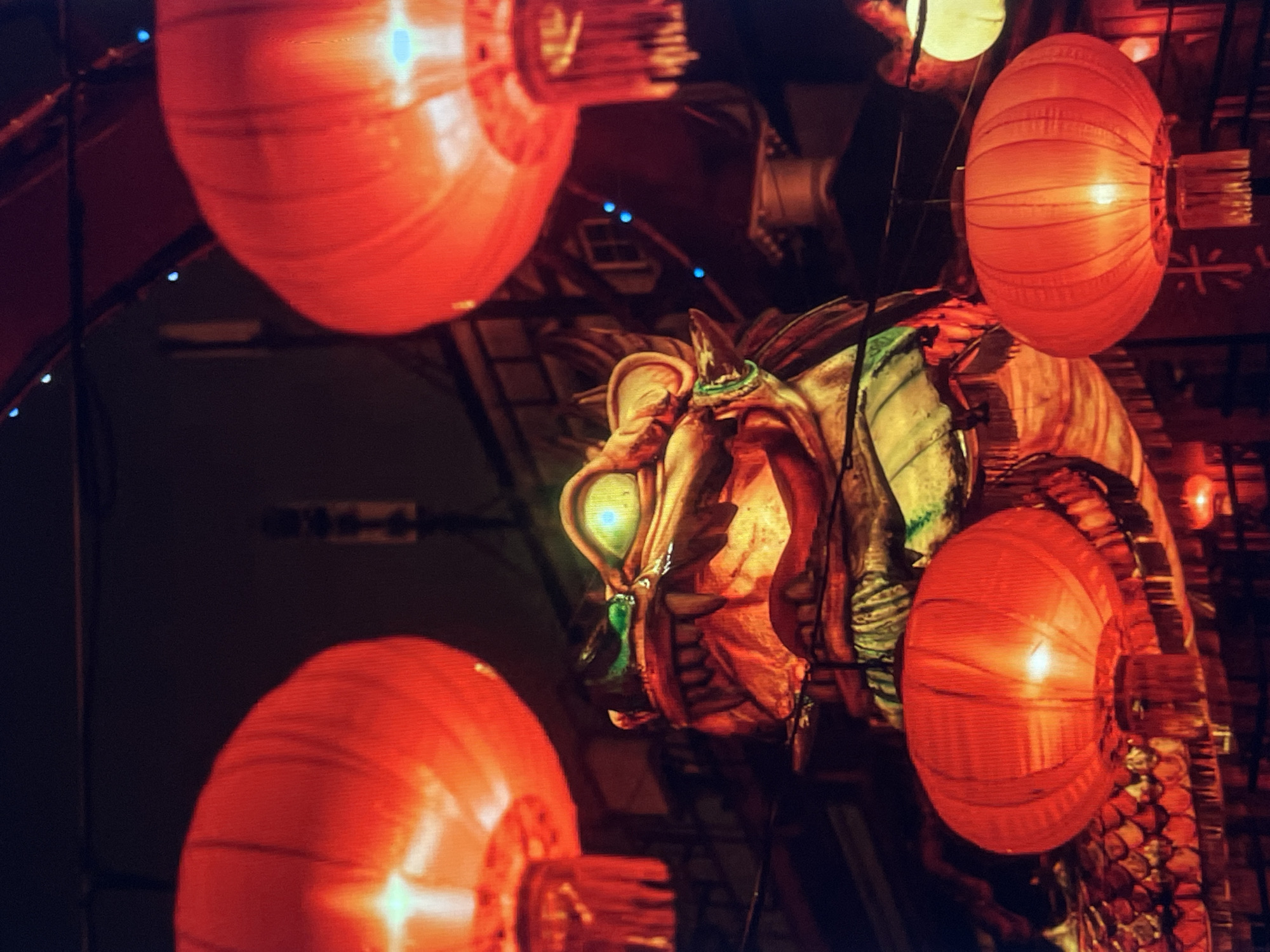 The image depicts a night scene featuring several illuminated red lanterns. A large, ornate dragon figure is centrally located, with its head and part of its body visible. The dragon has a light green eye and appears to be made of a textured material. The lanterns are of varying sizes and are positioned around the dragon. The background is dark and indistinct, with some hints of architectural elements and possibly other decorations. The location is unknown.