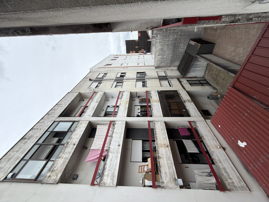 The image depicts a high-angle view into a narrow urban space between residential buildings in Bragança, Portugal, under an overcast sky. The foreground features a partial red corrugated metal roof.
To the left, a multi-story concrete apartment building exhibits weathered surfaces with peeling paint and exposed concrete. Its facade features several balconies with red metal railings.
* The uppermost visible balcony has a large glass window section above it and a pink fabric piece and a white, patterned fabric hanging over its railing.
* The middle balcony on the left displays a wooden rocking chair, a white box-like object, and a brown cardboard box. A white roller blind is partially lowered behind the railing.
* The lowest visible balcony on the left has white and pink laundry hanging over its railing and additional white laundry items suspended on a line further back.
Centered in the mid-ground, another multi-story building, lighter in color, is visible. It also features balconies with red railings, one of which has a small satellite dish attached. Laundry is visible hanging on multiple balconies of this building. A vertical drainpipe runs down its facade.
The ground level, mostly obscured by the foreground roof, reveals a concrete-paved courtyard area. Two small, dark-roofed structures, resembling sheds, are situated in this area. A dark-colored waste bin stands near the base of the center building's wall. A tall, textured concrete wall, part of an adjacent building, frames the right side of the image, showing similar signs of wear. Beyond the courtyard, in the background, another building with a dark, possibly tiled roof is partially visible. No individuals are present in the frame.