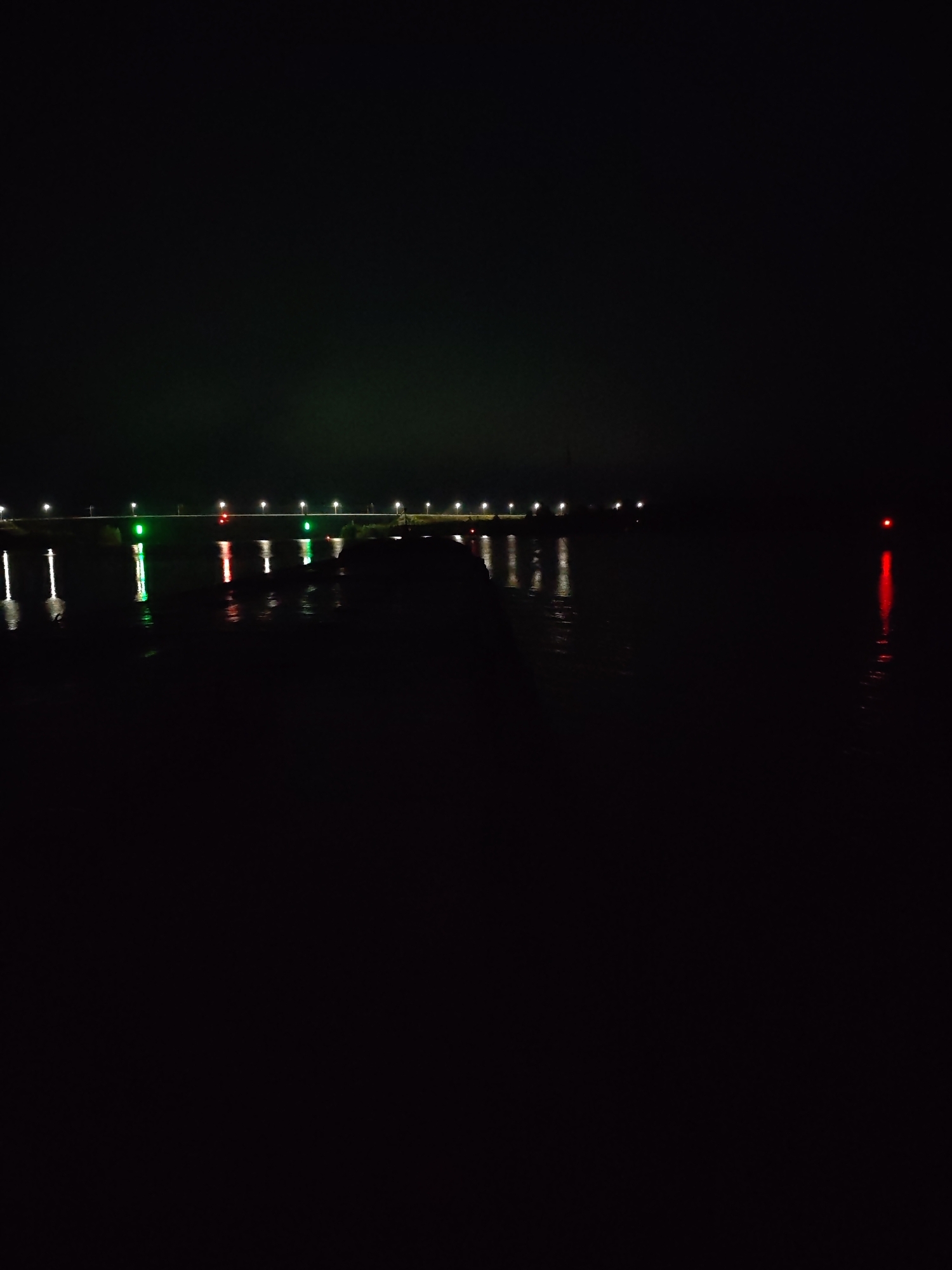 This is a nighttime photograph of a body of water, likely a river or lake, with a bridge or pier in the background. The scene is dark, indicating it is well after sunset. Lights are visible along the length of the bridge, some appearing as white streetlights and others as green and red navigational lights. These lights are reflected in the dark water, creating streaks of color. The water itself is calm, with minimal ripples. To the far right of the image, a solitary red light is visible, also reflected in the water. No people are visible in the image. The overall mood is serene and quiet, typical of a nighttime scene by the water. The location context suggests this scene might be in Ivanov Bor, Russia.