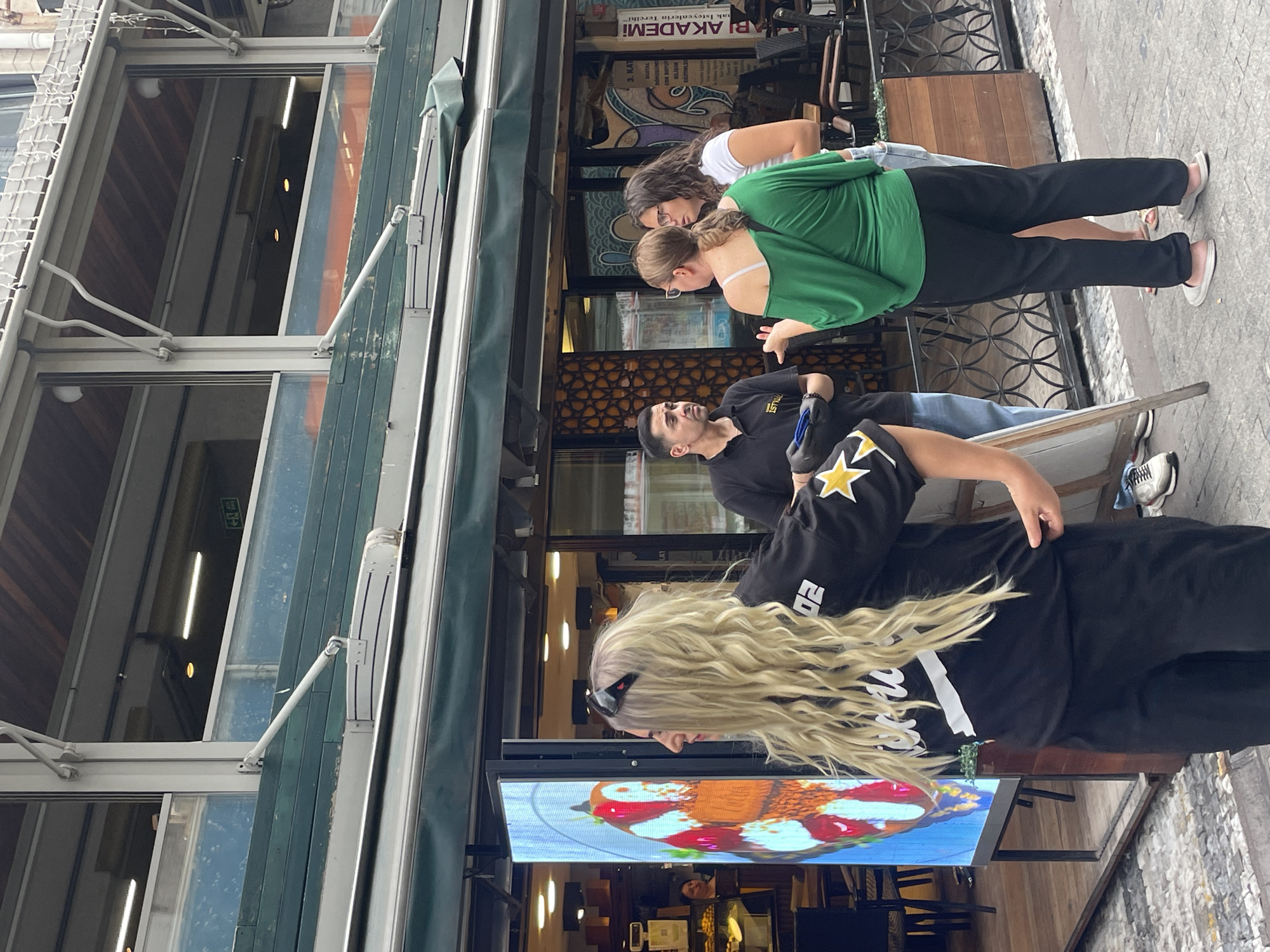 In Kadıköy, Turkey, on a cobblestone street, a group of four people are gathered in front of a restaurant with green awnings. A woman with long blonde curly hair, wearing a black t-shirt with a yellow star and white lettering, and black pants, stands in the foreground, facing left. To her right, a man in a black polo shirt and jeans holds a blue object. He is speaking with two women, one wearing a green off-the-shoulder top and black pants, and the other in a white t-shirt and light-colored shorts. The restaurant has large windows displaying various items and signage. An illuminated digital display stands to the left of the entrance, showing an image of colorful food. The overall scene depicts a casual street interaction in an urban setting.