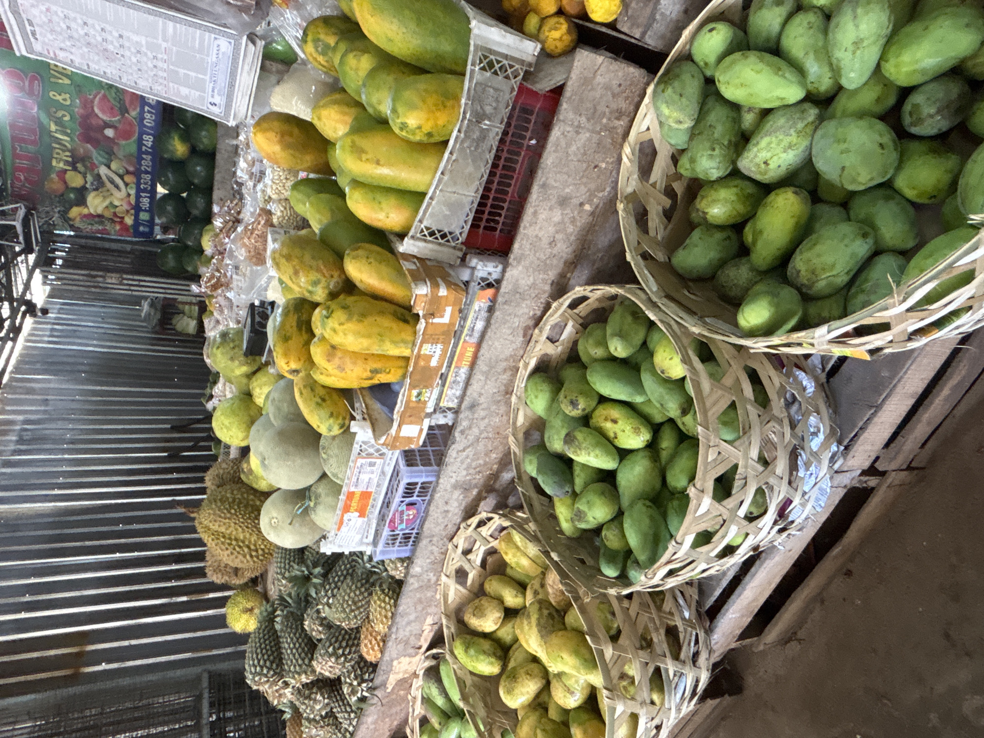 The image captures a vibrant fruit market scene, possibly in Ungasan, Indonesia, judging by the Southeast Asian fruits and the phone number on the sign. The market is filled with an abundance of fruits, including pineapples, melons, durian, papayas, and what appear to be green mangoes or similar tropical fruits stacked high in woven baskets. The scene is lit by natural light, suggesting an outdoor or semi-outdoor setting. The market appears busy, with fruits neatly arranged for sale. The fruits are the main subject, filling most of the frame, highlighting their variety and freshness. The scene is captured from an angled perspective, possibly by someone standing slightly above the fruit display.
