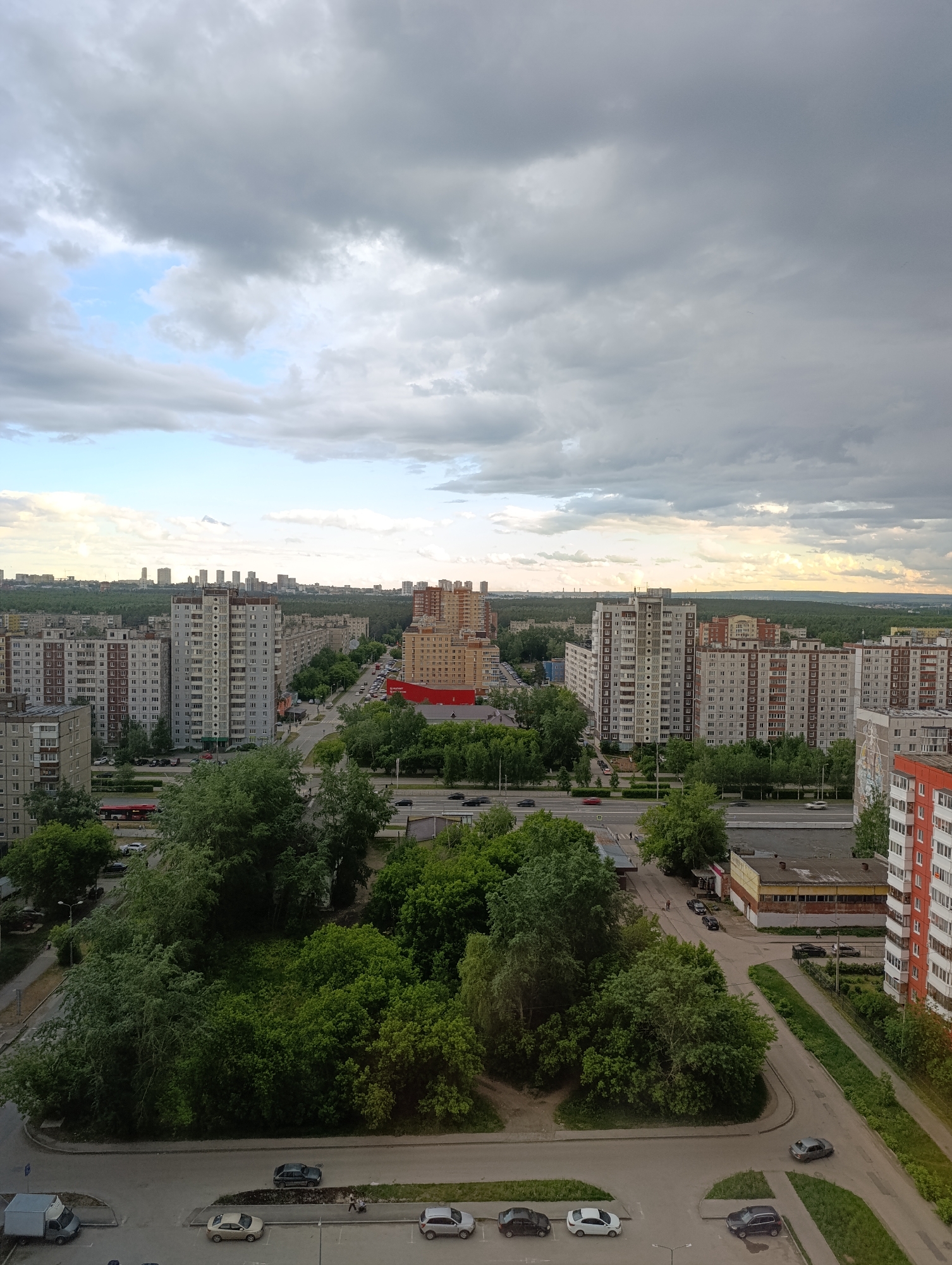 The image shows a high-angle view of a residential area in the city of Perm, Russia. Several multi-story apartment buildings dominate the landscape, arranged in rows and clusters. The buildings are constructed with concrete panels, typical of Soviet-era architecture. A large red building stands out among the grey and beige tones.
Streets and roads intersect the residential blocks, showing moderate vehicular traffic, including cars and a red bus. Green spaces, consisting of trees and bushes, separate the buildings and line the roads, adding patches of green to the urban setting.
In the foreground, a parking area is visible with several parked cars. Two people are walking on the sidewalk. The sky is overcast with heavy clouds, casting a soft, diffused light over the scene. A small patch of blue sky is visible through the clouds.