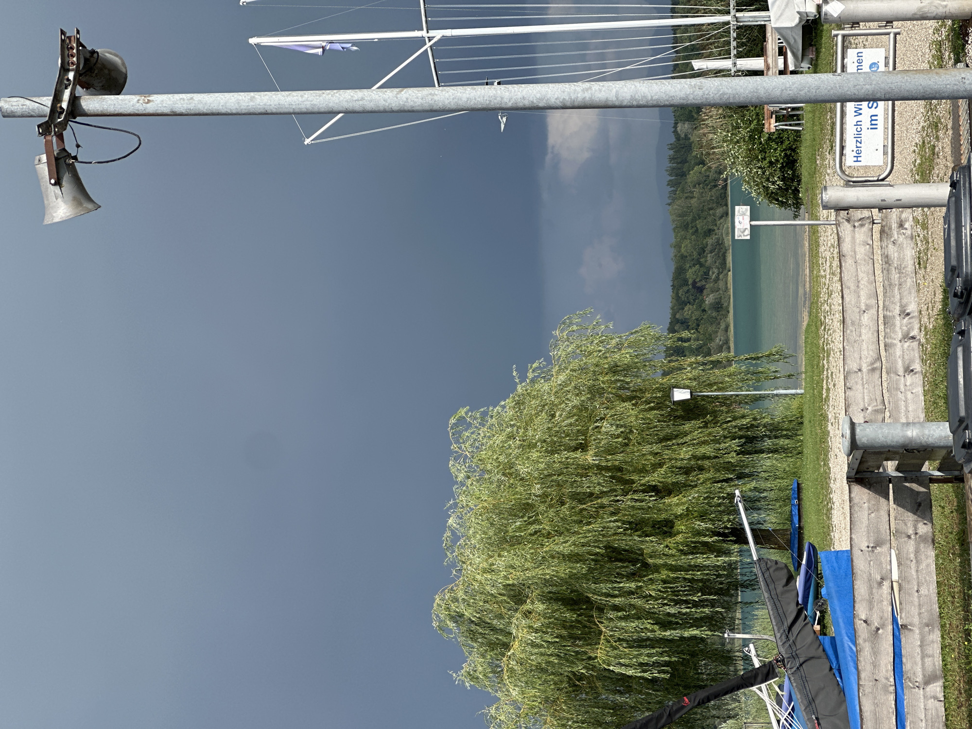 This image depicts a lakeside scene in Chiemsee, Germany, under a dark, cloudy sky. In the foreground, a weathered wooden fence runs horizontally across the frame. To the right of the fence, a metal gate with a sign reads "Herzlich Willkommen im S" (Welcome to the S...). Behind the fence, several sailboats or dinghies are covered with blue tarps, and a large weeping willow tree dominates the mid-ground. A lamppost stands near the tree, and beyond that, a clear turquoise lake stretches towards a tree-lined shore and distant hills. On the right side of the image, a tall metal pole supports a large loudspeaker and a smaller siren or bell. The sky is overcast with dark storm clouds gathering.