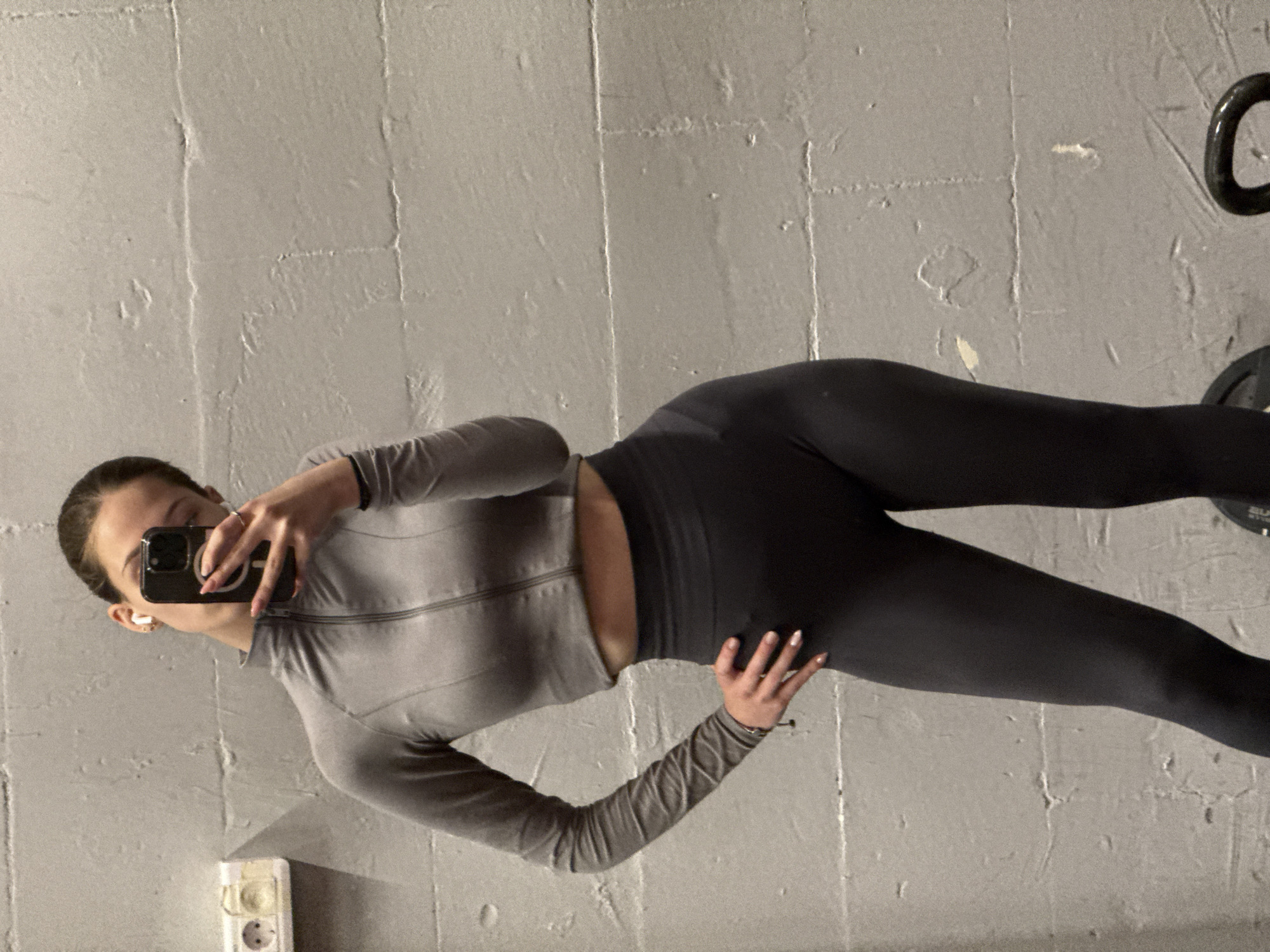 A person is taking a selfie in front of a wall. The person is wearing grey athletic clothing. In the background is a weight and a wall. The location cannot be determined from the image.