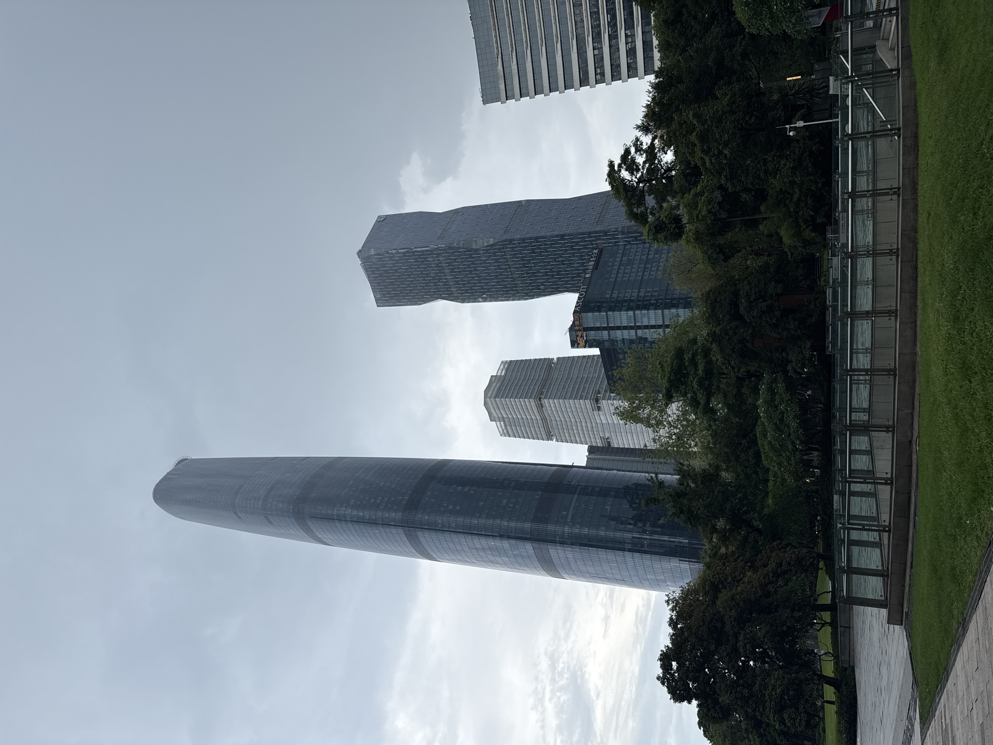 The image shows a panoramic view of modern skyscrapers in the Tianhe District of Guangzhou, China, under an overcast sky. The foreground features a well-maintained green lawn and a paved walkway. In the mid-ground, lush green trees form a dense line, partially obscuring lower levels of the buildings. A glass-paneled barrier with metal railings runs across the mid-ground, suggesting a park or public space.
Dominating the scene are several tall, contemporary buildings. The most prominent is a massive, sleek, dark-glass skyscraper with a distinctive curved, tapering form, leaning slightly to the left. To its right, another skyscraper stands, characterized by its angular, twisted design with a dark facade. Further to the right, a rectangular, light-colored building with numerous horizontal window bands is visible. Other buildings, including one with the "ASCOTT" logo, are also present, contributing to the urban skyline. The overall impression is one of a bustling, modern city district characterized by impressive architecture. There are no people visible in the image.