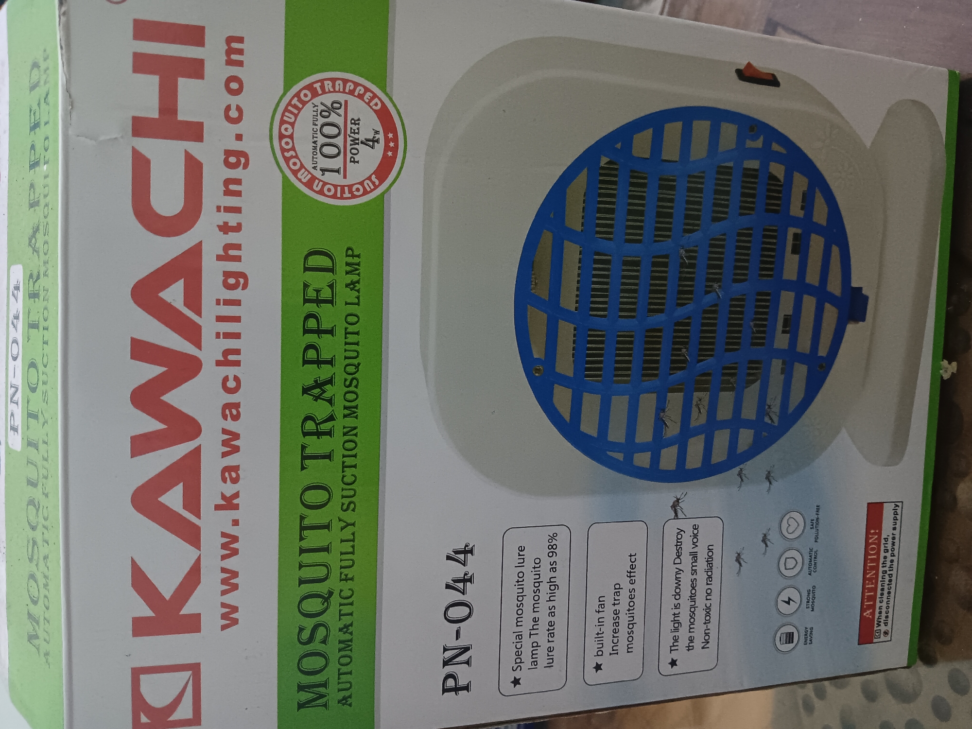 This is a product image of a "KAWACHI MOSQUITO TRAPPED" device, model "PN-044". The device is white with a blue grille and a red on/off switch. The packaging prominently displays the brand name "KAWACHI" and the product title "MOSQUITO TRAPPED AUTOMATIC FULLY SUCTION MOSQUITO LAMP". Key features are listed, including "Special mosquito lure lamp The mosquito lure rate as high as 98%", "built-in fan Increase trap mosquitoes effect", and "The light is downy Destroy the mosquitoes small voice Non-toxic no radiation". There are also icons indicating "ENERGY SAVING", "STRONG MOSQUITO", "AUTOMATIC CONTROL", and "SAFE POLLUTION-FREE". An "ATTENTION!" section advises to "When cleaning the grid, disconnect the power supply". The image appears to be a promotional photo taken in Batursari, Indonesia, likely for online retail or marketing purposes. The background suggests an indoor setting, possibly a home or retail space, with the product placed on a surface.