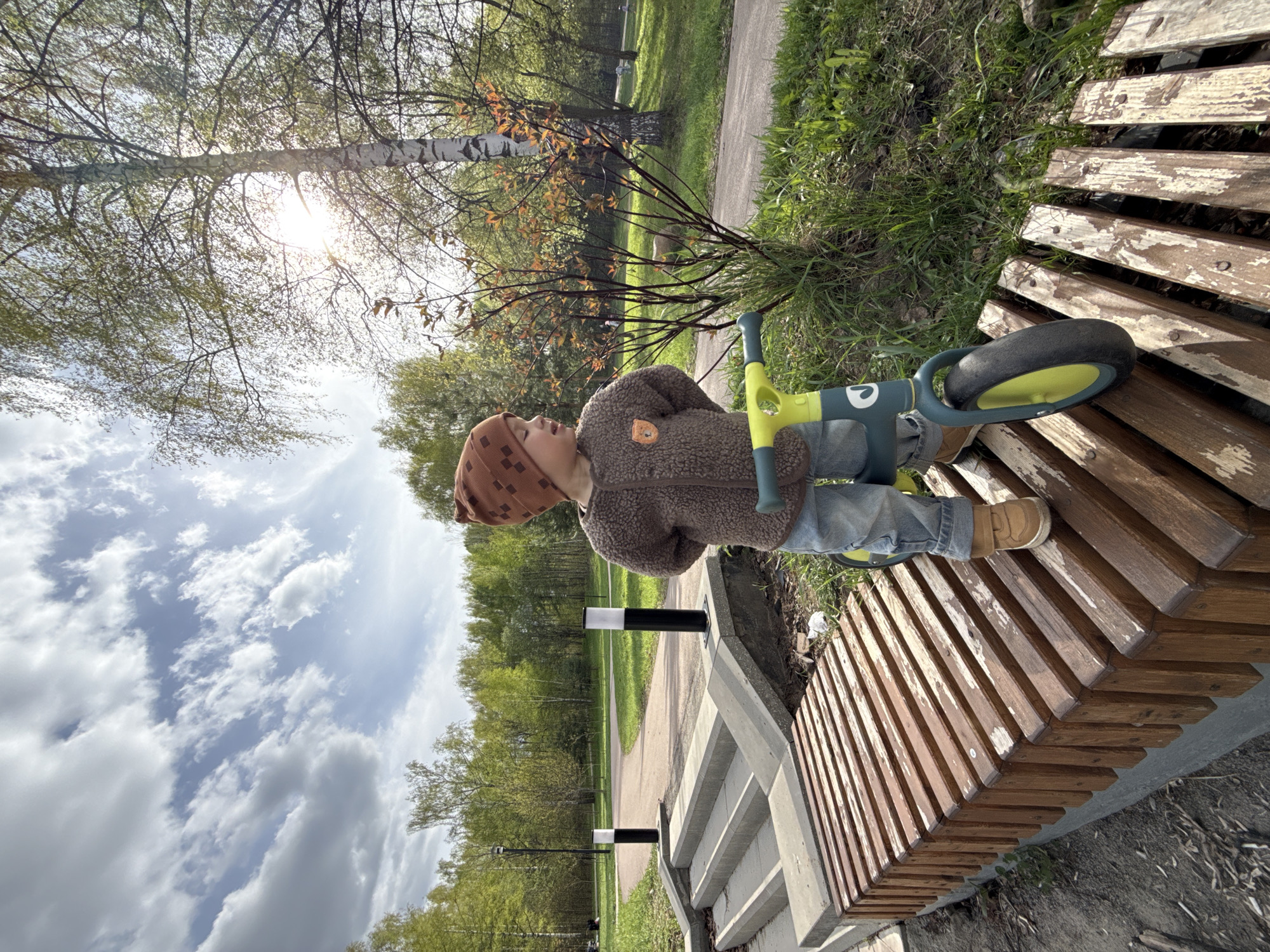 A child wearing a brown hat and jacket sits on a wooden bench with a green and yellow tricycle beside them. The bench is located in a park or green space, possibly in Rusanovo, Russia, indicated by the presence of deciduous trees and a walking path with black and white bollards. The sky is partly cloudy with sunlight filtering through the trees.