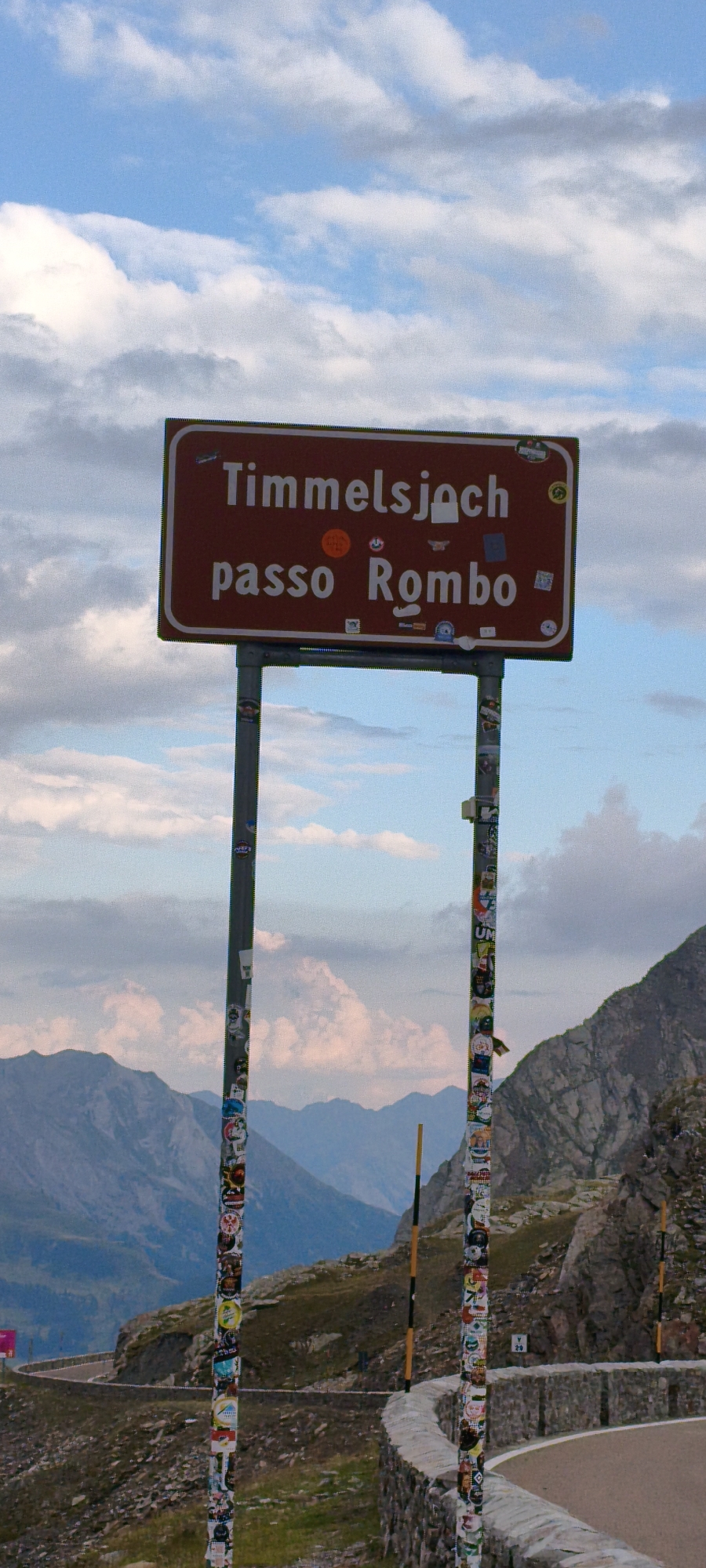 The image shows a road sign indicating "Timmelsjoch passo Rombo." The sign is a rectangular, dark red sign with white lettering, mounted on two metal poles. The poles are covered in numerous colorful stickers, suggesting it's a popular tourist spot. The sign is situated in a mountainous, outdoor environment. In the background, there are rugged, layered mountains under a cloudy sky with patches of blue. A winding road is visible to the left and right of the sign, with a stone wall bordering the road on the right. There are no people visible in the image. This location is the Timmelsjoch pass, which is the border between Austria and Italy. While Sölden is a town in Austria that is near the Timmelsjoch, the image itself does not provide specific indicators of Sölden itself, but rather the pass.