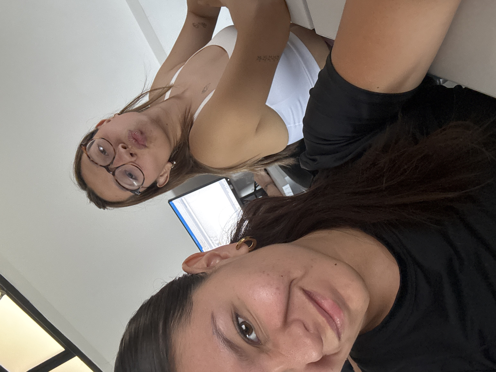 This media file features two women indoors, likely in San Andrés Cholula, Mexico, captured in a casual, low-angle selfie. The image is rotated counter-clockwise.
In the foreground, partially filling the lower right portion of the rotated image, is the face of one woman. She has dark hair, dark eyes, and a subtle smile, with a visible gold hoop earring on her ear. Her dark top is visible at her shoulder and neck.
Behind her, occupying the upper left of the rotated frame, is the second woman. She has light brown hair and is wearing tortoiseshell-framed glasses. Her lips are playfully puckered in a "duck face" expression. She wears a white tank top, revealing several small tattoos on her arms, including what appears to be a butterfly on one arm and a linear pattern of small stars on the other.
The setting is an indoor environment with light-colored walls or ceiling, suggesting a bright room. A computer monitor displaying what looks like a document or code with a blue scroll bar is partially visible in the background, hinting at an office or home office setup. The overall mood is relaxed and friendly. No explicit text is clearly legible in the image, apart from the unreadable content on the screen. The time of day is not discernible, as the lighting appears to be artificial or from an unseen window.