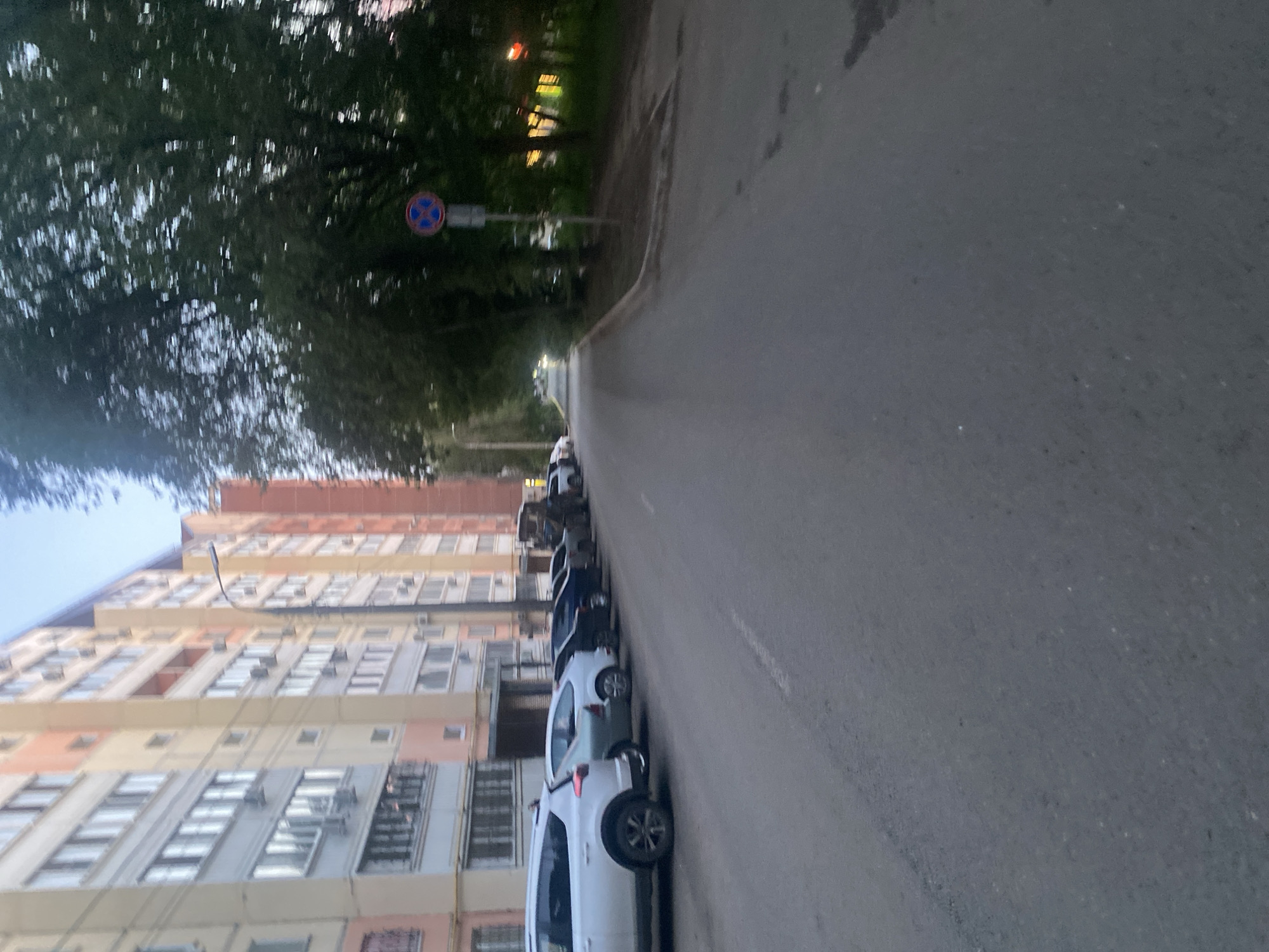 The image shows a street scene in the city of Samara, Russia. On the left, a multi-story apartment building with a light-colored facade lines the street. Several cars are parked along the curb in front of the building. On the right side of the street, there are trees and a street sign. The road appears to be paved and is in a state of disrepair, with cracks and patches visible. The lighting suggests it is either early morning or late evening.