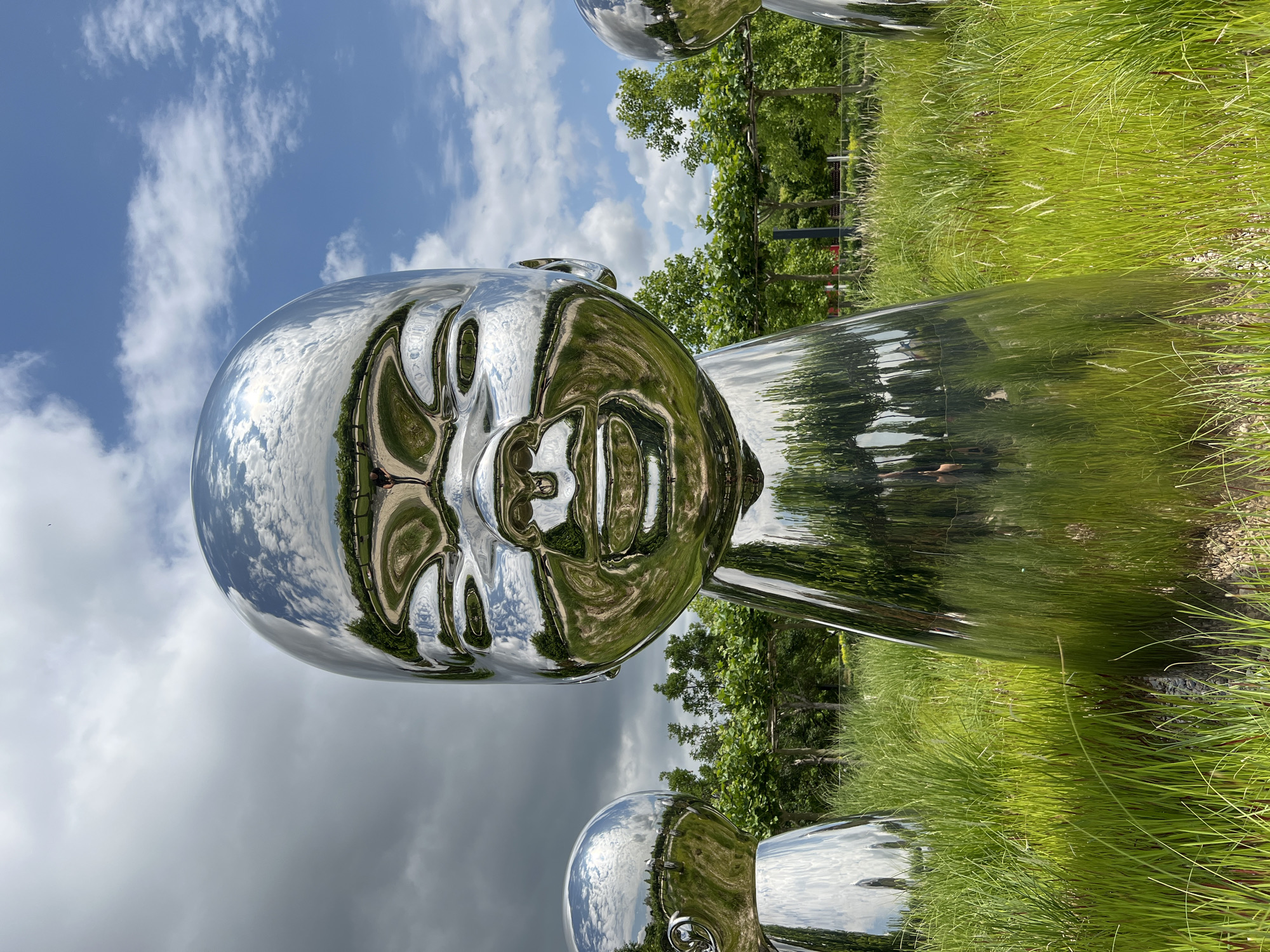 This image depicts a large, reflective sculpture of a baby's head, positioned in a grassy outdoor area. The sculpture is made of polished metal and reflects the surrounding environment, including trees, sky, and clouds. In the background, other similar sculptures are visible, as well as a pathway and trees. The sky is partly cloudy with a mix of blue and gray clouds. The foreground is dominated by lush green grass and a few exposed stones at the base of the sculpture. The overall scene suggests a park or art installation. This sculpture is located in Krasnodar, Russia.