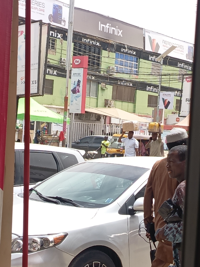 This image depicts a busy street scene in Fagge A, Nigeria, characterized by commercial activity. In the foreground, a silver sedan is parked, partially obscuring a darker sedan behind it. To the right, two men are visible: one wearing a brown garment and a light cap, partially obscured, and another man in a patterned top, carrying a dark object and looking to the right. In the midground, two men walk rightward, each balancing a cardboard box on their head. One wears a white shirt, the other a light brown shirt and is holding a phone to his ear. A person in a bright green shirt stands near a yellow tricycle further back.
The background features a multi-story building painted light green with black trim, extensively branded with "Infinix" logos and mobile phone advertisements. Other advertising banners for "itel," "CITY 100," "Note 40 Series," and "Android" are visible on poles and adjacent buildings. Overhead, numerous power lines crisscross the frame. A green tent structure is visible on the left side of the street, and a red vertical banner occupies the far left foreground. The scene suggests a vibrant urban commercial environment.