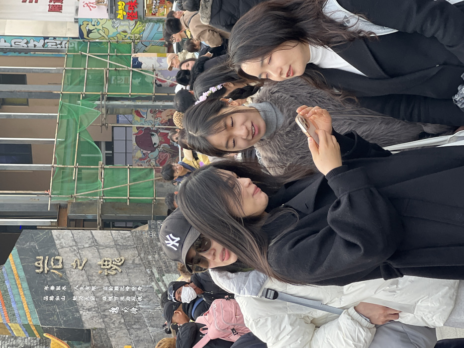 A group of people, mostly women and children, are gathered closely together, some looking at a phone. One woman is wearing a New York Yankees baseball cap and sunglasses. The background includes a stone monument with Chinese writing, scaffolding, and colorful building art. A sign with Japanese text is also visible, adding to the international feel of the scene. The crowd appears to be in a public space.