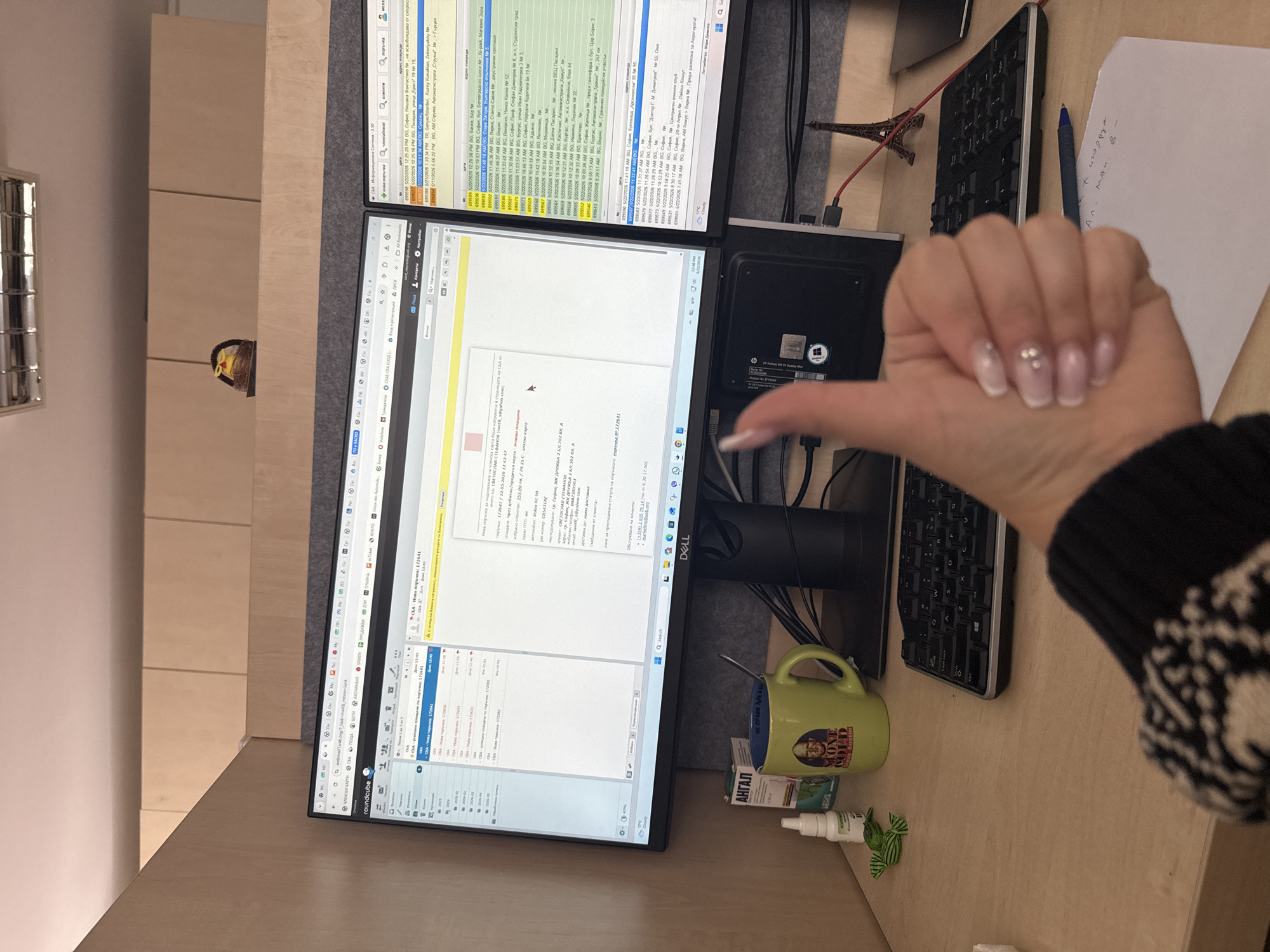 A medium-angle, indoor shot shows a desk with two computer monitors, a keyboard, and various objects, including a hand giving a thumbs-up. The left monitor displays email software, with a visible email about "СБА - Нова поръчка: 172641" (SBA - New Order: 172641). The right monitor displays a table of data. A green mug sits to the right of the bottom monitor, along with a bottle labeled "АНГАЛ." An Eiffel Tower figurine sits behind the monitors. The hand in the foreground has manicured nails. The desk is light brown, and a cabinet system is visible in the background. On the left monitor is a window with customer data. The OCR text indicates "София, ЖК ДРУЖБА 2" (Sofia, Druzhba 2 residential area) which indicates an address in Sofia, Bulgaria. The screen also shows addresses around Sofia.