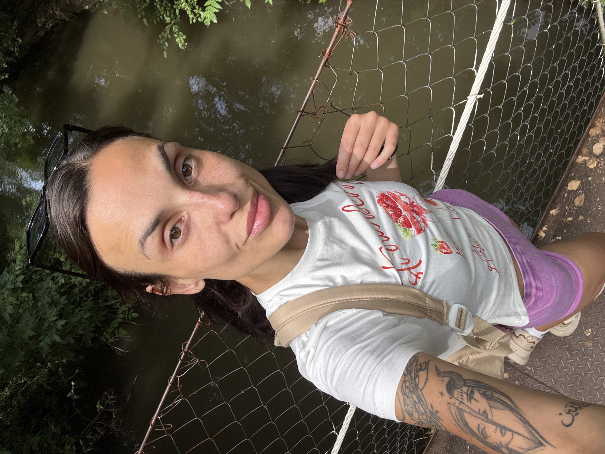 This is a selfie of a woman on a bridge overlooking a body of water. She is wearing a white t-shirt with a strawberry graphic and red text, pink shorts, and sneakers. She has a crossbody bag with a beige strap. A tattoo is visible on her right arm and leg. The scene appears to be outdoors, with greenery visible in the background. The time of day seems to be daytime, with sunlight casting shadows. The woman has a neutral expression. The bridge is constructed of metal with a wire mesh fence. The text on her shirt reads "Strawberry."
