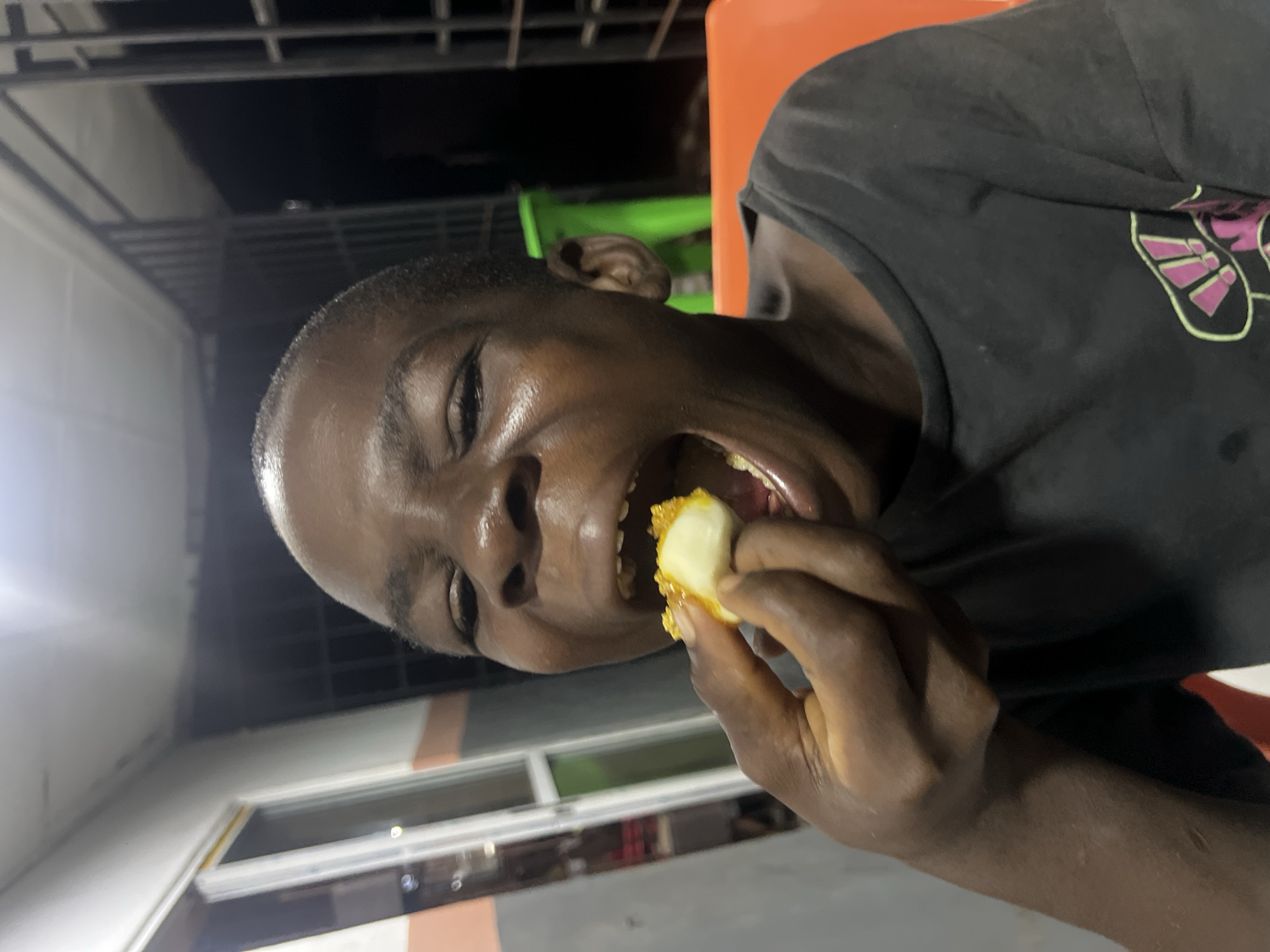 The image shows a close-up, sideways portrait of a young person with dark skin in Nkpor, Nigeria. The individual is actively eating, with their mouth wide open as they bring a piece of food to their mouth with their fingers. The food appears to be a light-colored, bite-sized item, partially covered in a textured, golden-brown coating, possibly indicating it is fried. The person's eyes are open wide, and their expression suggests enjoyment or anticipation of the meal. They are wearing a dark t-shirt, which has faint purple and lime green graphic elements visible on the shoulder area. The setting appears to be indoors or under a covered outdoor area, likely at night or in the evening, given the dark background and artificial light source illuminating the subject. Structural elements like ceiling beams or rafters are visible in the upper background, alongside hints of an orange object, possibly a chair, and a green item further back. No text is visible in the image.
