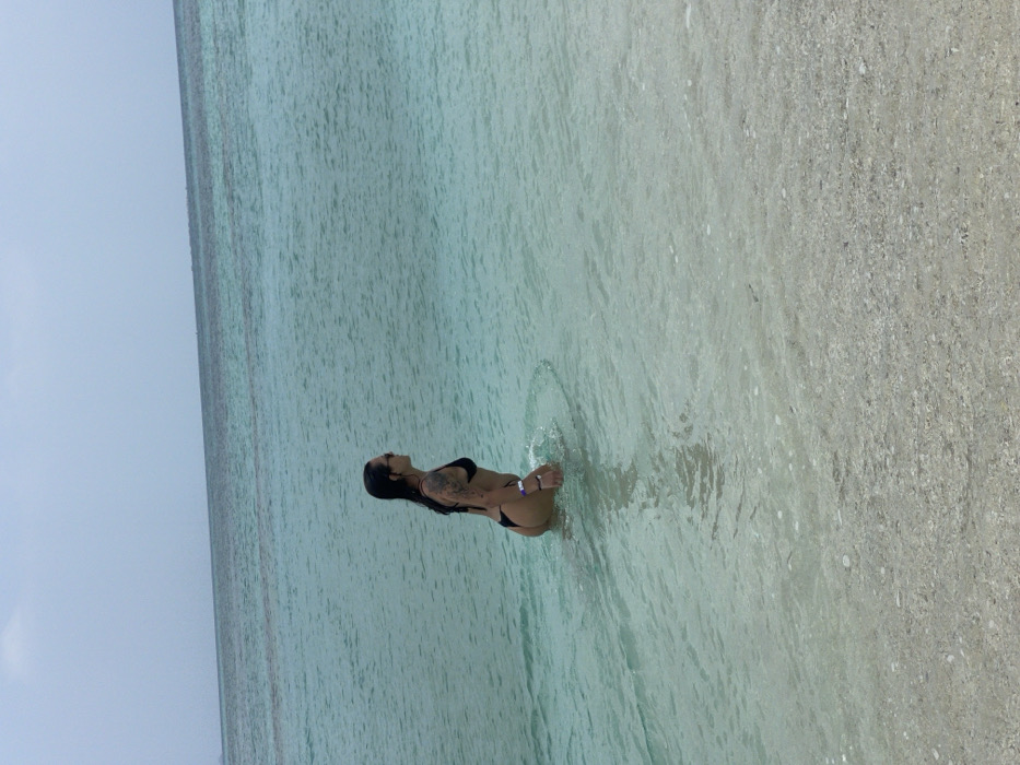 An adult female is depicted standing waist-deep in clear, shallow turquoise ocean water at a beach in Zanzibar, Tanzania. She is observed in profile, facing towards the right side of the frame. Her attire consists of a black two-piece bikini and sunglasses. Tattoos are visible on her left arm. Her hair is dark and appears wet. A distinct circular ripple pattern is evident in the water around her lower torso. The sandy seabed is visible beneath the translucent water in the foreground, transitioning to deeper blue ocean water towards the horizon. The sky above the horizon is hazy and light blue.
