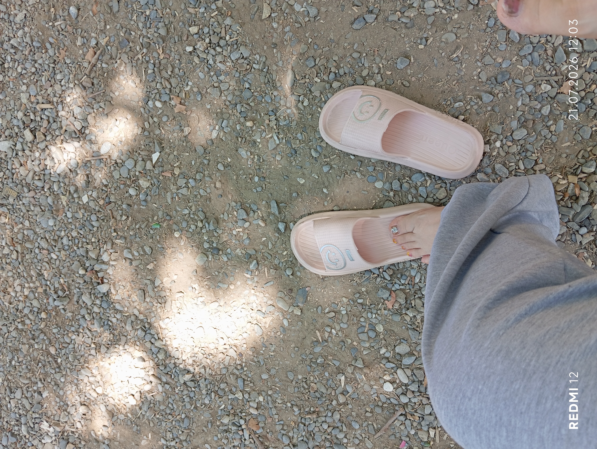 A person's feet, wearing light pink slides and grey sweatpants, are positioned on a gravelly surface. The slides appear to be made of a flexible material, with one featuring a small smiley face logo. The gravel is composed of small, grey and brown stones, with some scattered dry leaves and twigs. Sunlight casts dappled shadows on the ground, suggesting an outdoor setting, possibly in a park or a less-maintained area. The timestamp "21.07.2026 12:03" and "REDMI 12" are overlaid on the image, indicating the date and time of capture, and the device used. The location is identified as Bekabad, Uzbekistan.