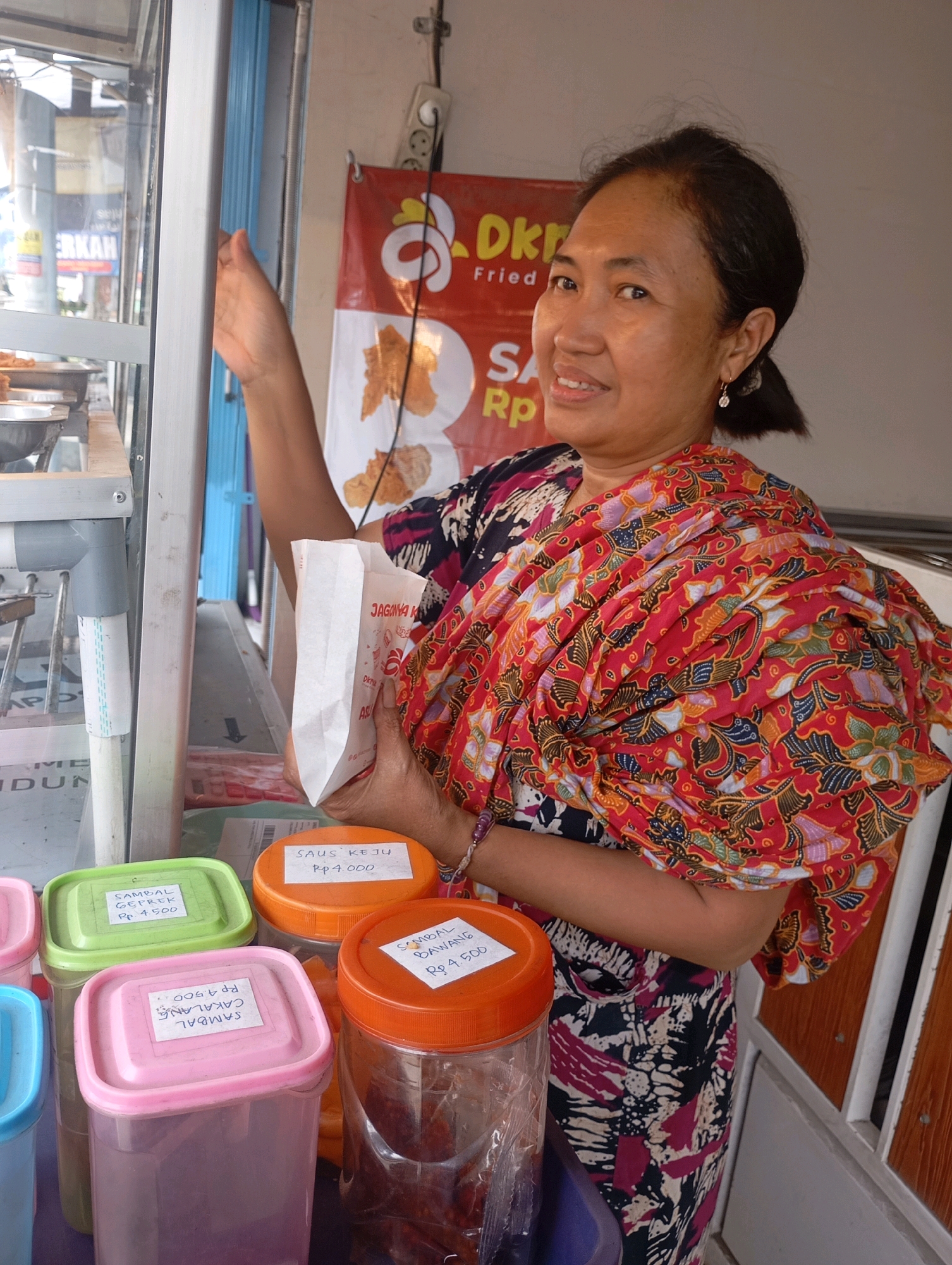 A middle-aged Indonesian woman with dark hair pulled back and wearing a colorful batik pattern outfit stands behind a food stall in Babelan, Indonesia. She is holding a white paper bag in her right hand and looking towards the camera with a slight smile. In front of her are several plastic containers of condiments, each labeled with their contents and price in Indonesian Rupiah. The stall appears to be selling fried food, indicated by a banner in the background that reads "DKR Fried Chicken". The overall setting is a street-side food vendor, common in Indonesian cities.