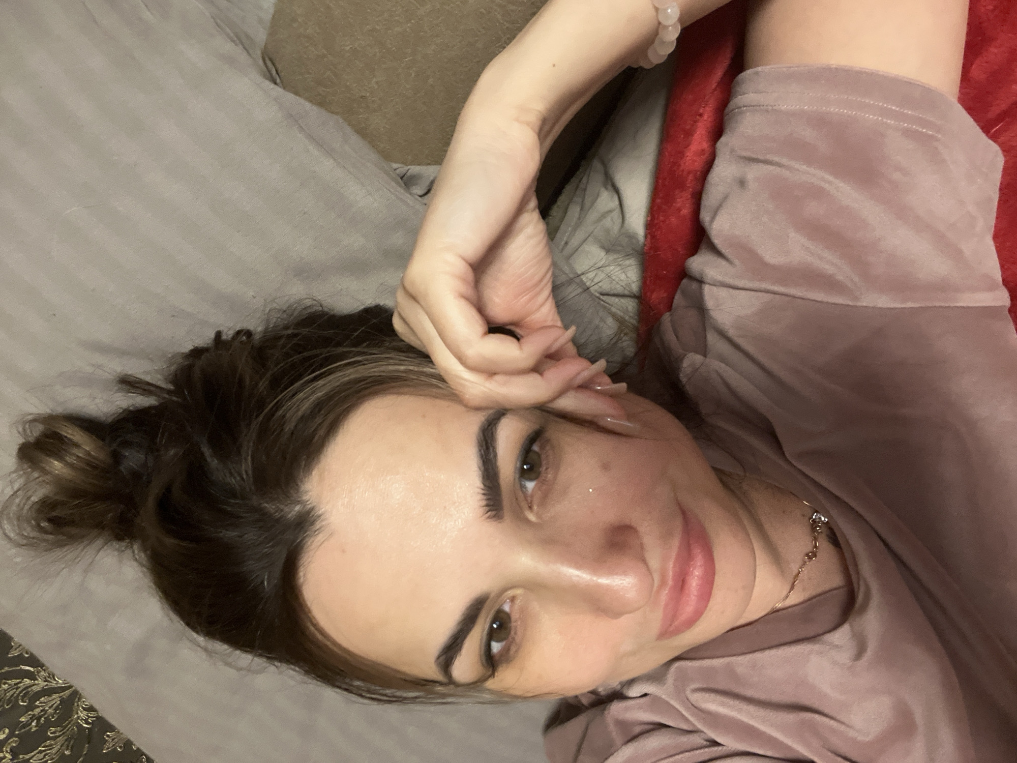 A woman is reclining on a bed, possibly indoors, in a location of Unknown, Unknown. She is dressed in a pink, velvet-like top, with a bead bracelet on her left wrist. Her hair is pulled back into a bun, and she is wearing a delicate necklace. Her left hand is raised towards her forehead. She is lying on a gray pillow and a red blanket.