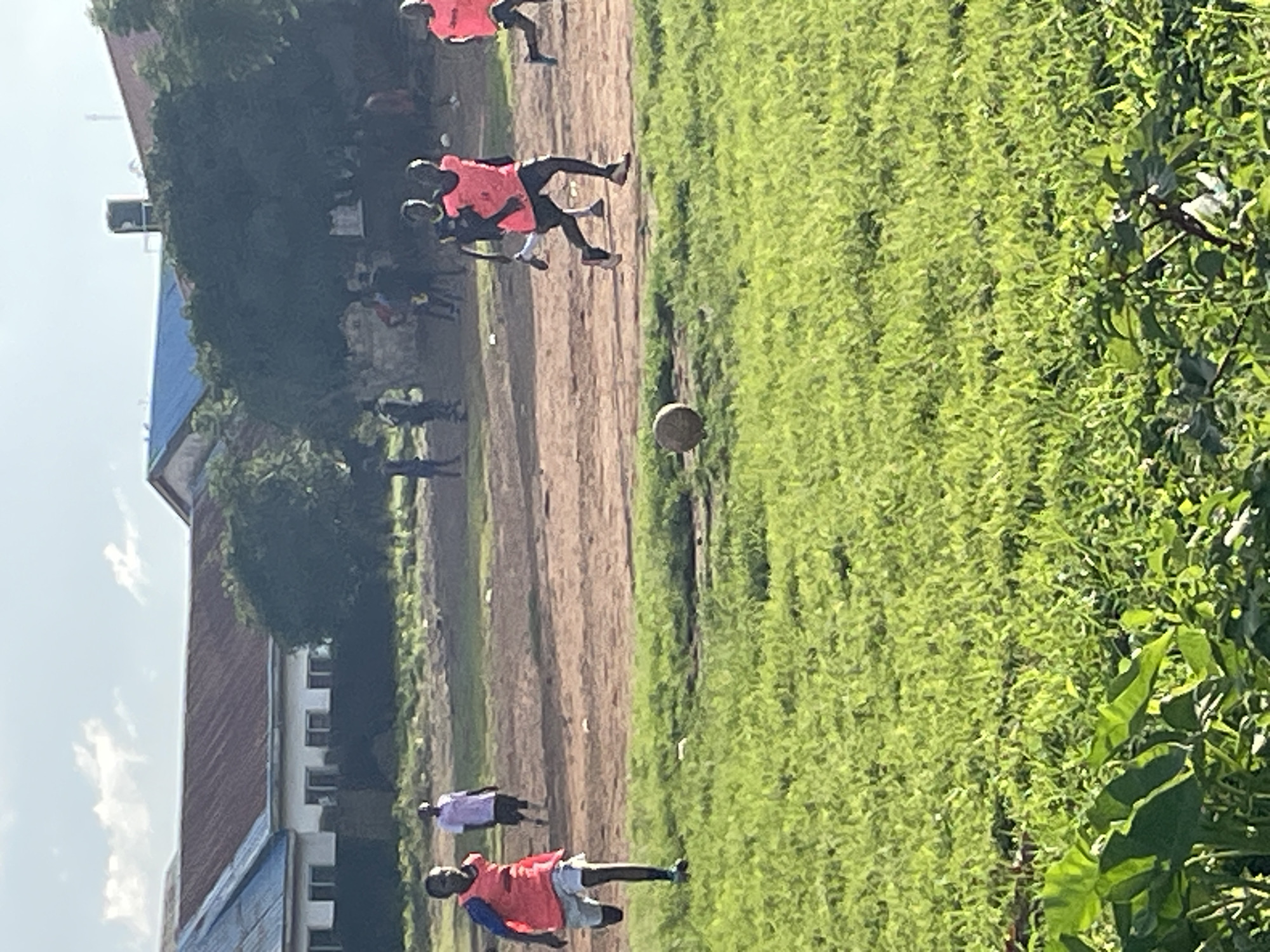 A group of young boys are playing soccer on a dusty field next to a grassy area. The boys are wearing brightly colored vests over their t-shirts, with some wearing pink and others yellow. One boy in a pink vest is running towards the viewer with the ball at his feet. Another boy in a pink vest is further down the field, also running. In the background, there are buildings with red and blue roofs, and trees. A water tower is visible behind one of the buildings. The sky is partly cloudy. The scene appears to be in Jos, Nigeria, a city known for its soccer culture. The image captures a moment of casual recreation, highlighting the passion for the sport among the youth in the region.
