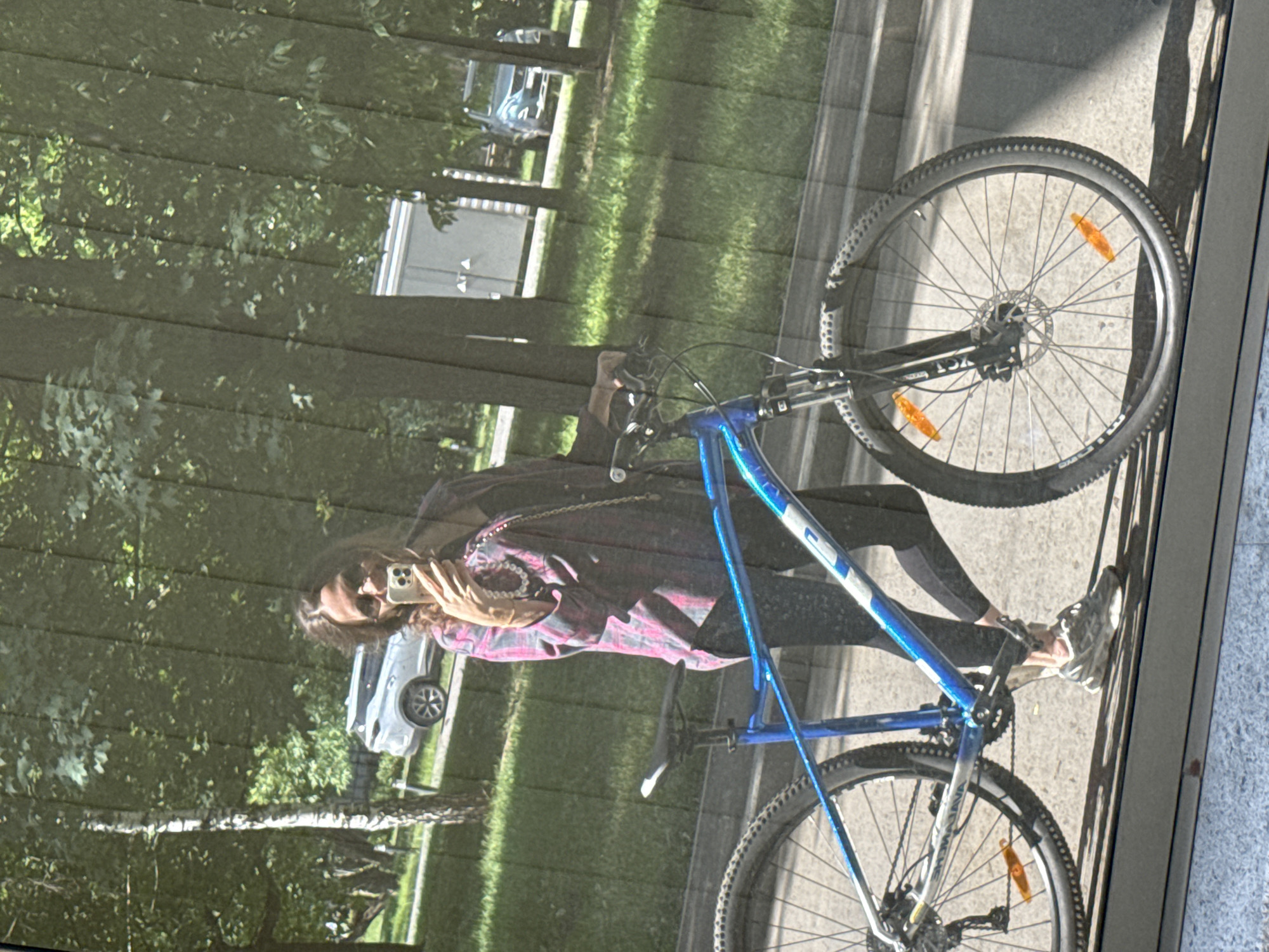 The media file shows a reflection of a person taking a selfie with a bicycle. The person is wearing a pink and black plaid shirt and dark leggings, with white athletic shoes. They are holding a smartphone in their right hand to take the picture. The bicycle is blue and black, with a silver frame.
The scene appears to be outdoors, with trees and grass visible in the background, suggesting a park or a street lined with trees. There are also reflections of buildings, including what looks like a white structure. A silver car is also reflected in the background.
The lighting suggests it is daytime, likely sunny given the shadows. The overall mood seems casual and relaxed, with the person seemingly enjoying their time outdoors.
The image is a reflection on a glass surface, which distorts the scene slightly and adds a layer of visual interest. The photograph is taken vertically, with the main subject rotated, indicating the camera was likely held in portrait mode.
No visible text is decipherable in the image. The location is specified as Moscow, Russia.