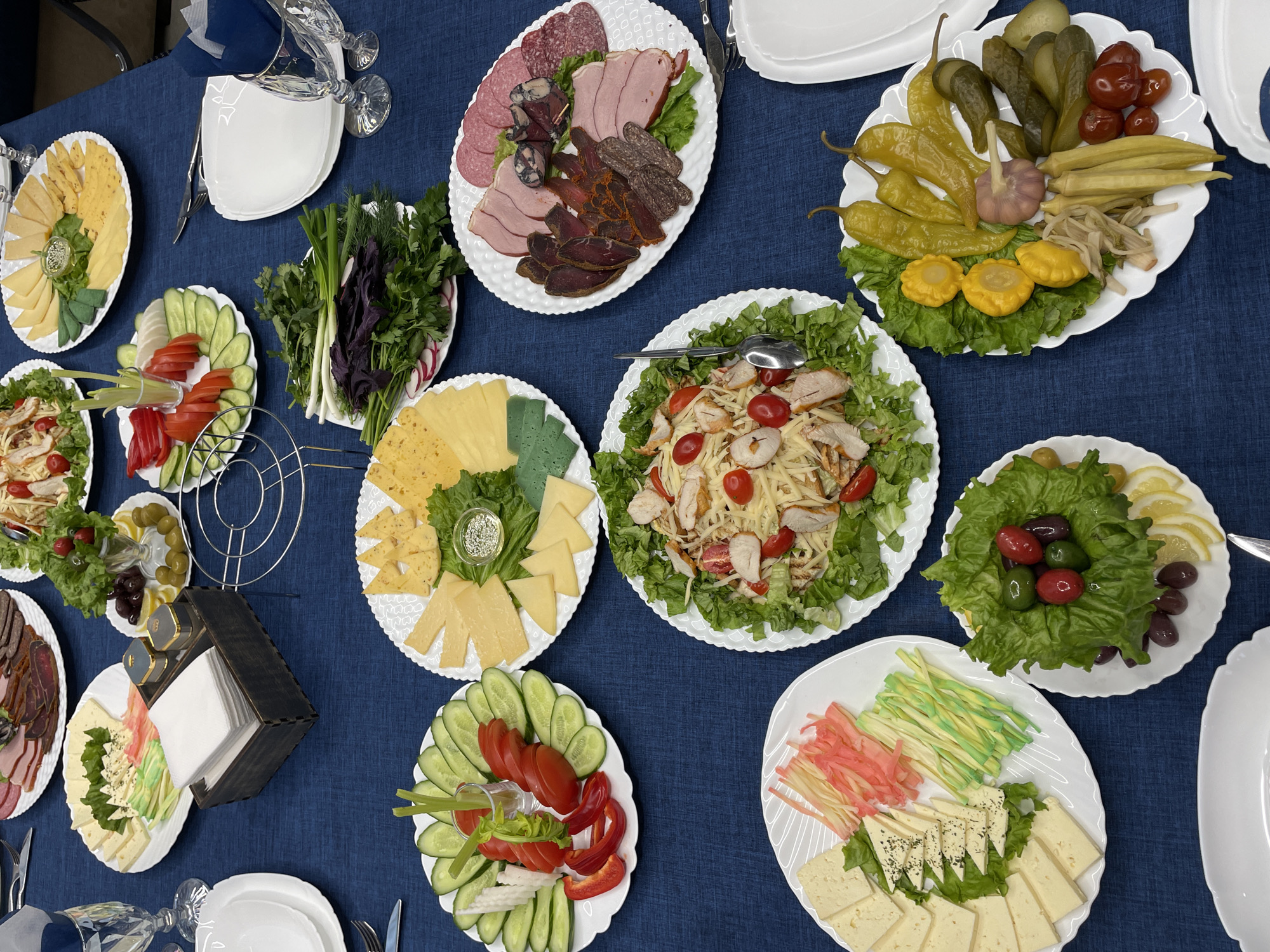 An overhead shot captures a richly laid table covered with a dark blue tablecloth, prepared for a gathering in Solontsy, Russia. The table is laden with a variety of dishes, arranged on white, often scalloped-edged, plates.
The subjects are diverse food items presented in a buffet style. Several plates feature an assortment of cheeses, including yellow, light yellow, and green varieties, some arranged with fresh lettuce and herbs. Cold cuts are prominently displayed on one large plate, showcasing salami, ham, cured meats like basturma and sujuk, all on a bed of lettuce. Another large plate holds a selection of pickled vegetables, including cucumbers, cherry tomatoes, banana peppers, garlic, pattypan squash, okra, and what appears to be pickled cabbage.
Two different salads are visible: one with shredded chicken or shrimp, cherry tomatoes, and grated cheese over lettuce, and another with whole olives (green and black), red cherry tomatoes, and lemon slices nestled in lettuce cups. Fresh vegetable platters include sliced cucumbers, tomatoes, red bell peppers, radishes, green onions, dill, and parsley. One unique plate features thinly cut pink and green strips (possibly fish or vegetable) alongside white cheese with black specks, possibly herbs.
Other objects on the table include several empty white plates, clear drinking glasses with ornate stems, silverware (forks, knives, spoons), and a dark wooden napkin holder containing white napkins, flanked by small salt and pepper shakers. A metal stand, possibly for a tiered dish, is also visible.
The scene depicts a festive or celebratory meal, suggesting an event like a party or banquet. There are no people visible, allowing the focus to remain on the meticulously prepared food. The lighting suggests an indoor setting, likely during the day or evening with artificial light. No visible text is present within the image itself.