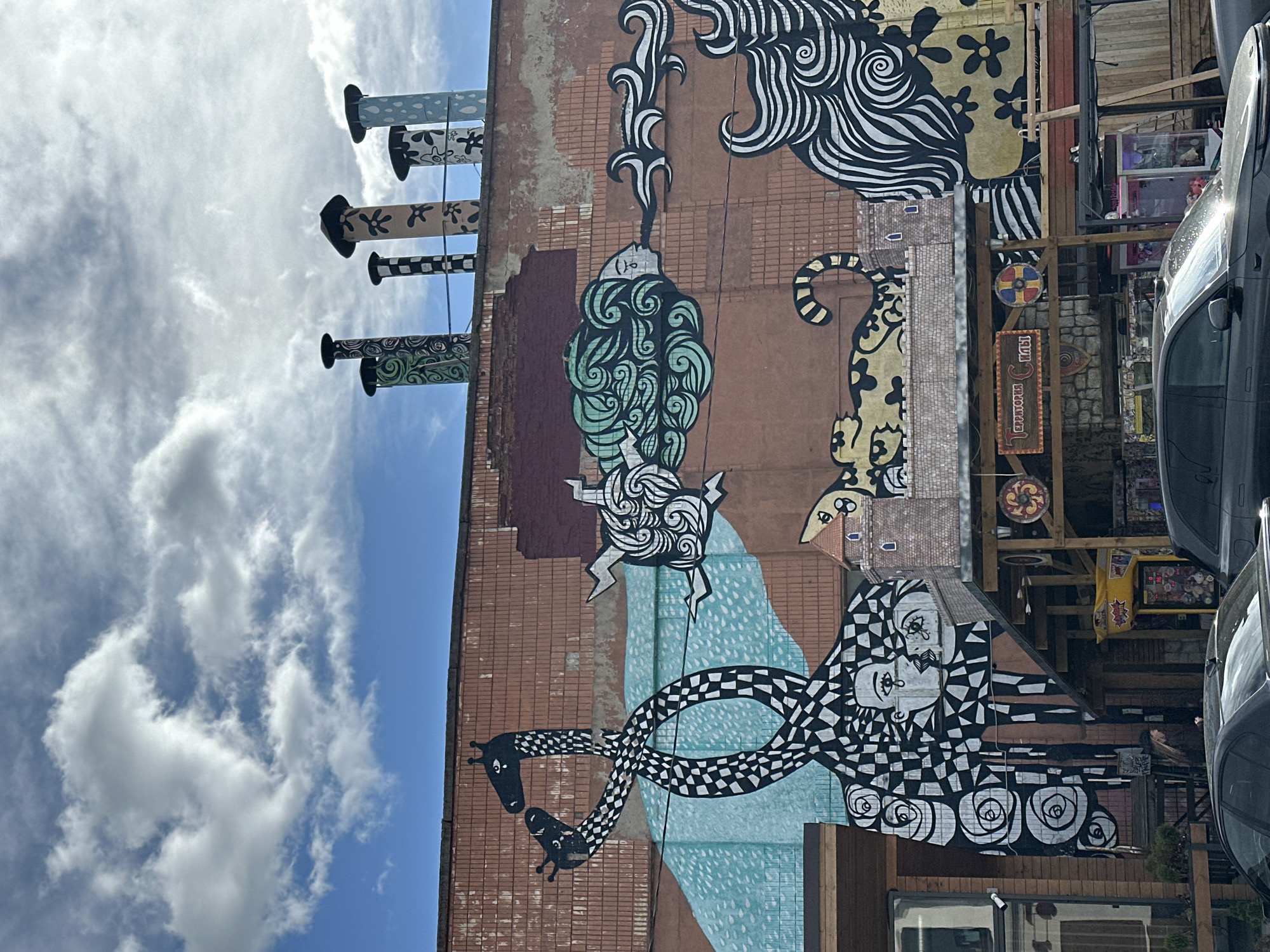 Here is a description of the image:
The image depicts a large mural painted on the side of a brick building. The mural features a variety of whimsical and abstract designs, including stylized animals, faces, and swirling patterns. The color palette includes brown, black, white, green, and light blue. Several cylindrical structures, possibly chimneys or vents, protrude from the top of the building and are painted with decorative patterns. The sky, visible to the left of the building, is blue with scattered clouds. The building appears to be part of a commercial area, with parts of storefronts and vehicles visible on the right side of the image. Based on the mural's distinctive style and the information provided in the search query, the image is likely taken in Minsk, Belarus.