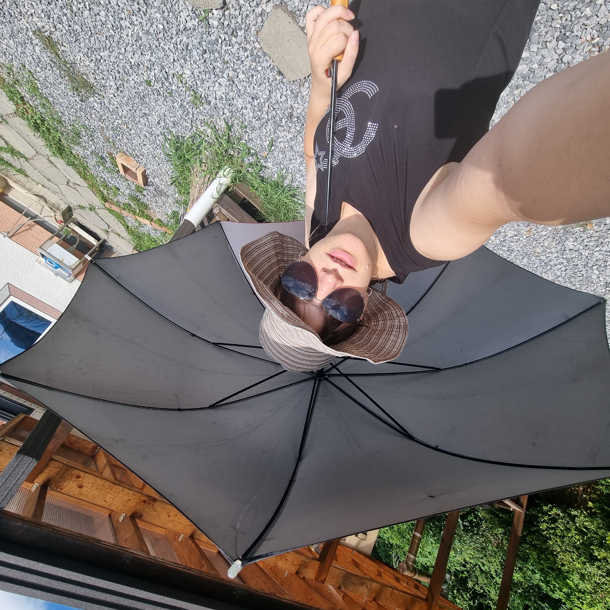 In Dunnae-myeon, South Korea, a person is taking a selfie outdoors. They are holding a large, gray umbrella over their head, shielding them from the sun. The person is wearing a black tank top with a sparkling logo and a wide-brimmed, striped hat. They are also wearing large sunglasses that reflect the sky. The background shows a gravel area, some green vegetation, and a wooden structure with a translucent roof. A white downspout is visible near the person. The scene suggests a sunny day, possibly in a backyard or garden setting.
