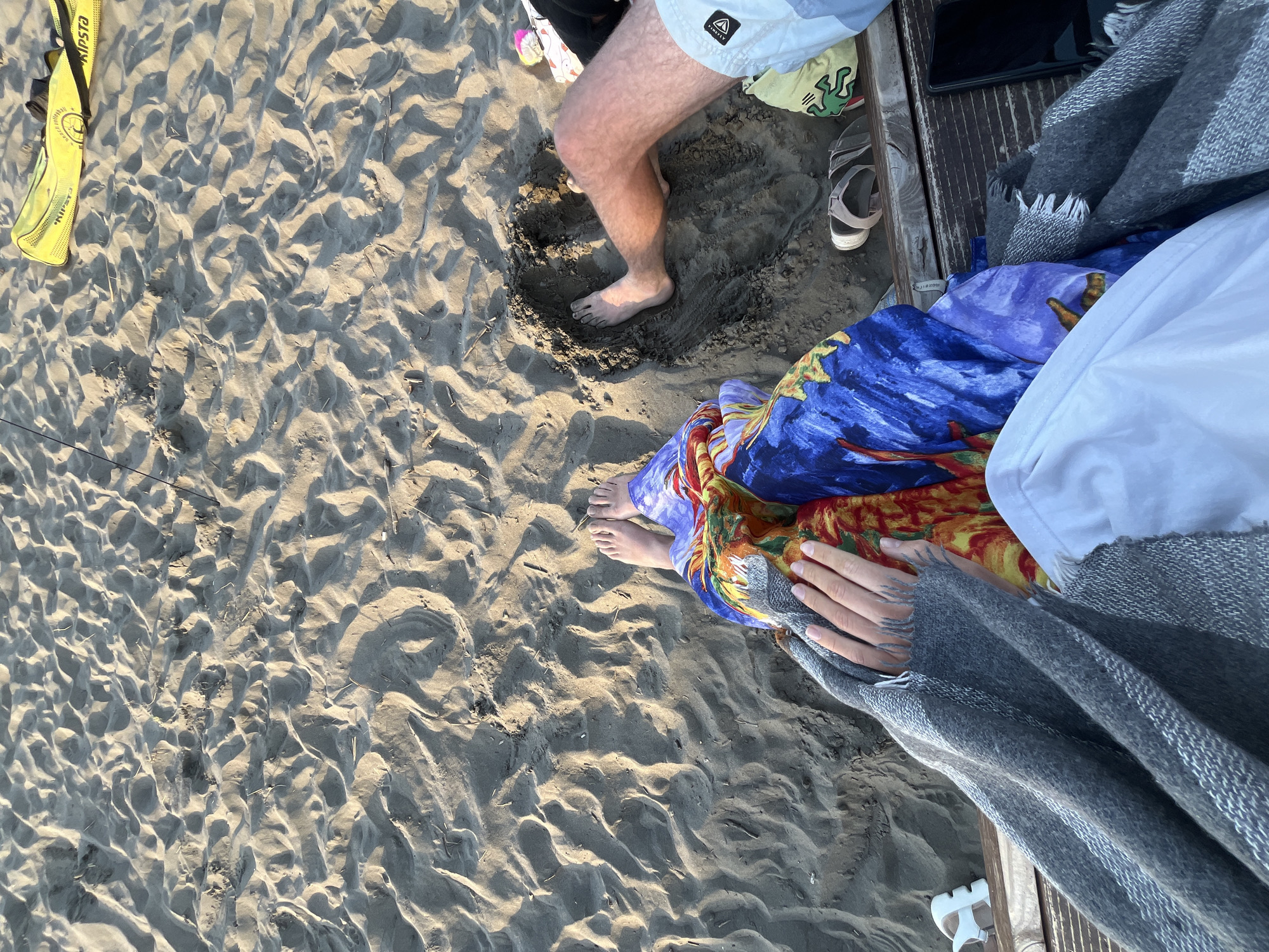 The image shows a top-down view of a sandy beach in Pistula, Montenegro. Two people are present, partially visible. One individual's legs are extended, with their right foot in a dug-out sand hole. This person is wearing light-colored swim shorts. The other individual's legs are bent, covered by a colorful patterned fabric and a grey blanket. Their hands are visible, one with tattoos.
The beach is covered in rippled sand, suggesting wind or water movement. Scattered on the sand are a yellow Kipsta beach volleyball bag, a pair of flip-flops, and a white sandal. A wooden structure, possibly part of a bench or boardwalk, runs horizontally across the lower portion of the image. A black smartphone is placed on this structure to the right. The lighting suggests it is late afternoon or early evening.