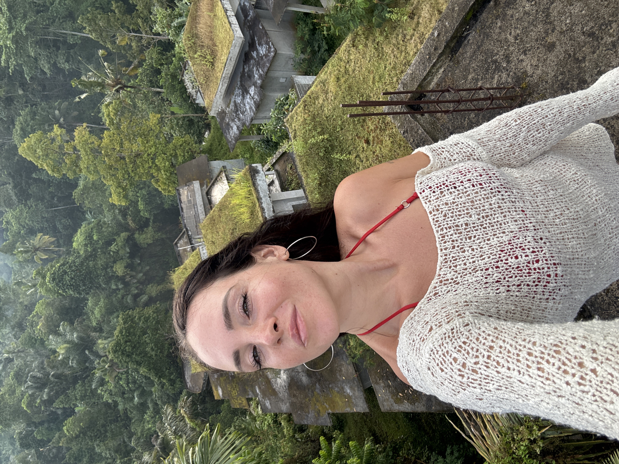 A woman with long, dark hair and fair skin is taking a selfie from a high vantage point, looking down and to her left with a slight smile. She is wearing a white, open-knit sweater over a red bikini top. Large hoop earrings are visible. The background reveals a lush, green jungle landscape with several abandoned, concrete buildings. These structures feature flat roofs covered in moss and vegetation. The overall scene appears to be in Bebandem, Indonesia, given the dense tropical foliage and the style of architecture, suggesting an overgrown, remote location.