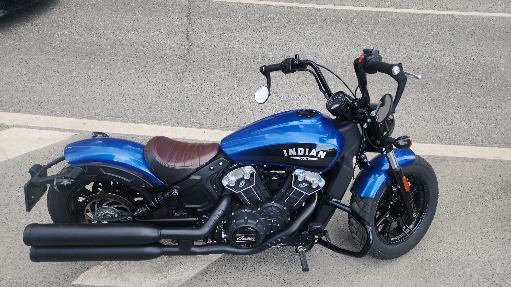 The image shows a blue Indian Scout motorcycle parked on an asphalt surface. The motorcycle has a brown leather seat, black handlebars, and a black exhaust system. The scene appears to be an outdoor setting on a street or parking lot, as the ground shows asphalt with some line markings. There are no people visible in the image. It is not possible to determine the location (Moscow, Russia) based on the visual information available.