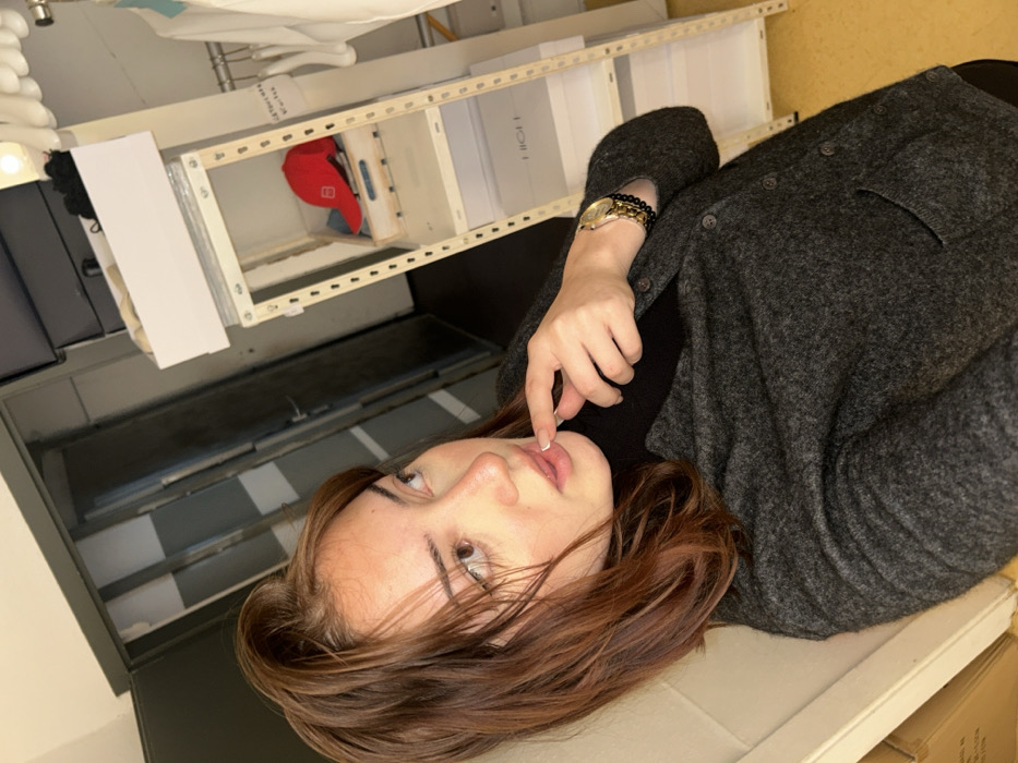 An indoor photograph depicts a female subject in a dressing room or storage area in Strasbourg, France. The individual, with medium brown hair, is wearing a dark gray button-down cardigan over a black turtleneck. On her left wrist, she wears a gold-toned watch and a dark beaded bracelet. Her right index finger is positioned at her lower lip, and her gaze is directed upwards and to her left. The background on the left features a partially open dark-colored door. To the right, a white metal shelving unit holds a white box containing dark objects on its top shelf. A lower shelf displays a wooden crate holding a red baseball cap. Below this, two white shoeboxes are visible, one explicitly labeled "HIGH." A clothing rack with white hangers and white garments is partially visible to the far right. The floor is yellow. No specific landmark or precise address within Strasbourg is discernible from the visual content.