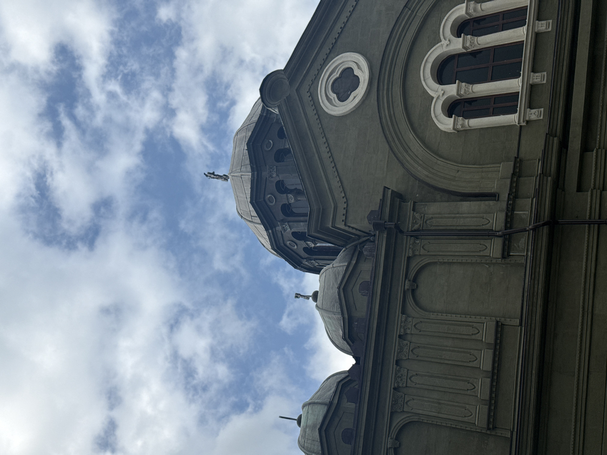 The image shows the facade of a building, likely a church or religious institution, under a cloudy sky. The architecture features several domes, with a prominent central dome topped with a cross. The building is constructed of stone or concrete and has decorative elements such as arched windows, columns, and ornate trim. The color palette is dominated by shades of green and gray, with the sky displaying patches of blue and white clouds. The perspective is from a low angle, looking up at the building. While the specific location is not explicitly identified within the image itself, given that the request specifies the city of Burgas, Bulgaria, this building is likely a significant religious structure within that city, such as the St. Bogoroditsa Church. There are no people or animals visible in the frame.