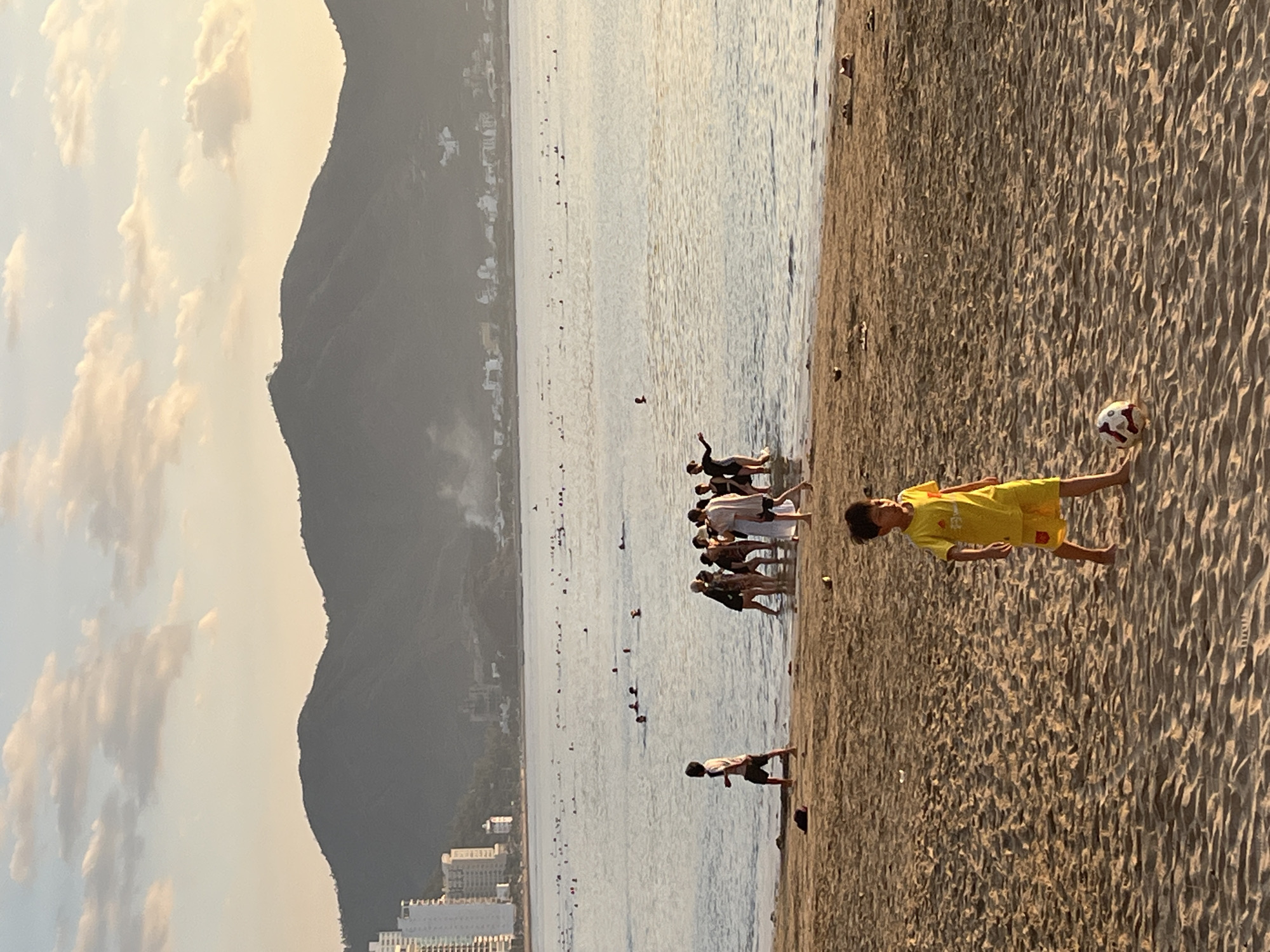 The image shows a beach scene in Bac Nha Trang Ward, Vietnam, at sunset. The beach consists of light brown sand. A group of people are standing in the water near the shoreline, some knee-deep. Closer to the foreground, a child wearing a yellow shirt and shorts is positioned on the sand next to a soccer ball. Another person is walking parallel to the shoreline and is knee-deep in the water. In the background, a mountain and a coastline with numerous buildings are visible. The sky contains clouds.