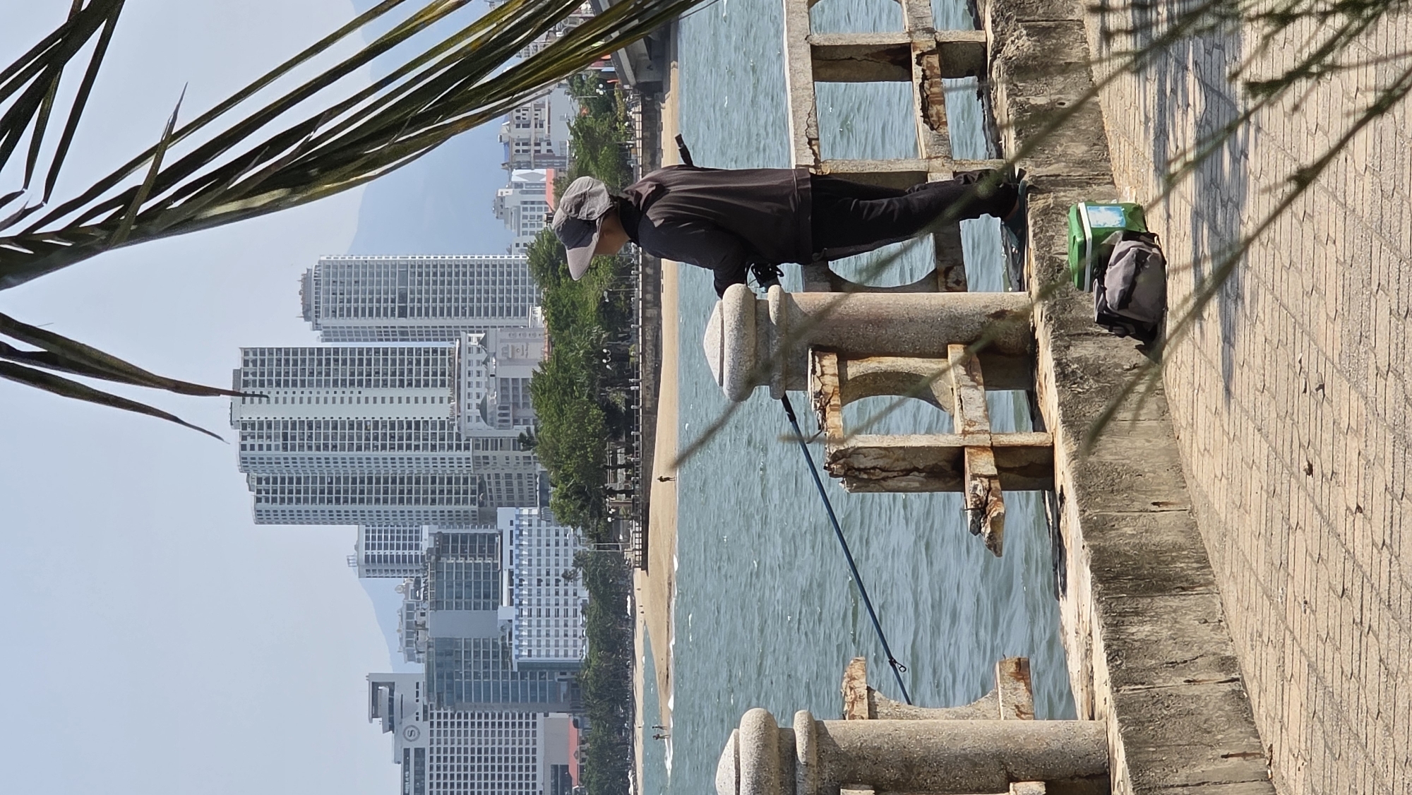 An image shows a man fishing off a concrete structure with the coastal city of Bắc Nha Trang, Vietnam, in the background. The man is wearing dark clothing and a light-colored hat. He is holding a fishing rod that extends into the blue ocean. Beside him is a small green bucket and a gray backpack on the side of the structure. The structure appears to be an old pier or part of a bridge. The backdrop features several high-rise buildings along the sandy beach. Palm fronds are visible on the upper-left corner of the image. The weather is sunny with clear skies.