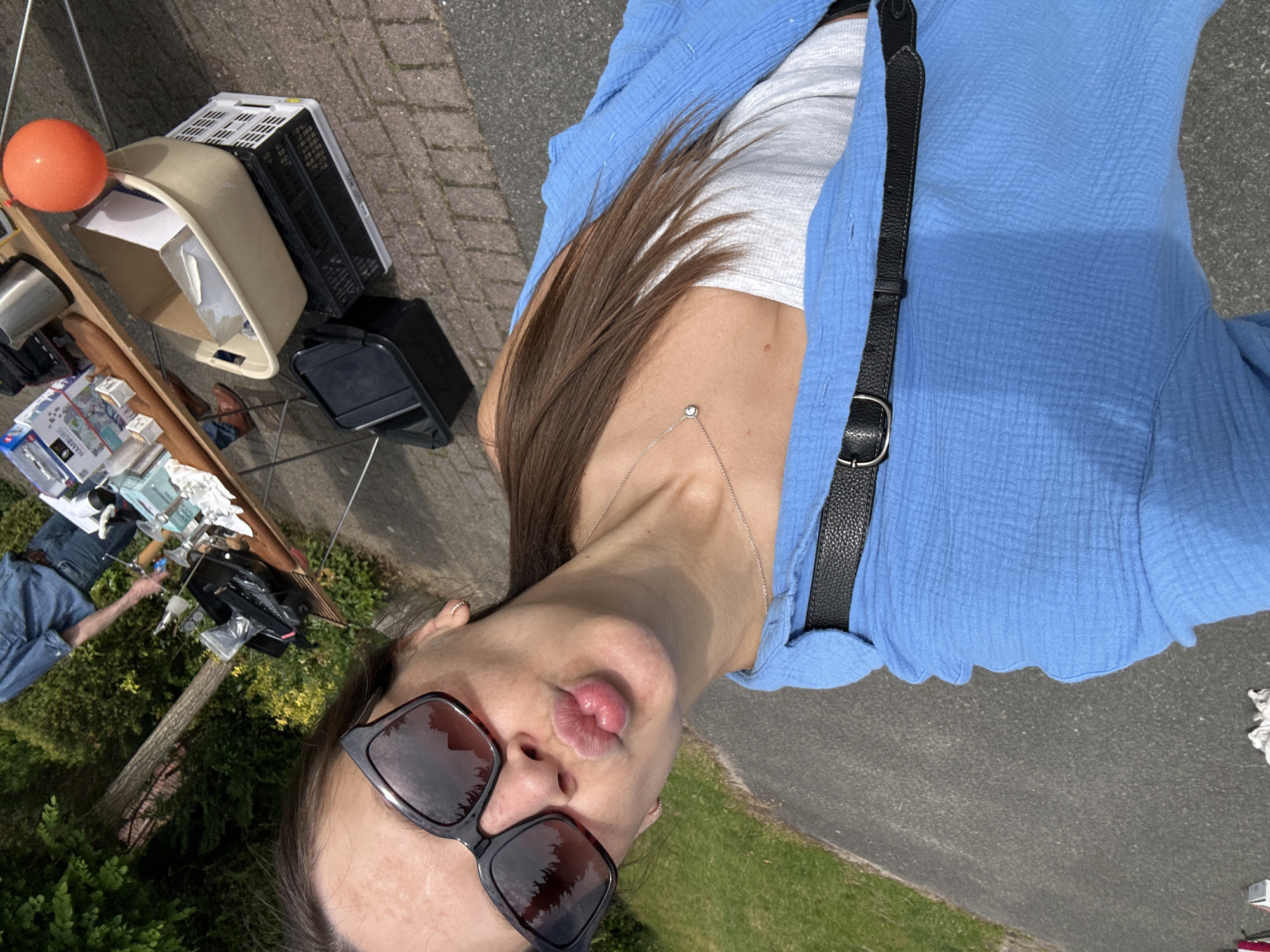 A selfie-style photograph shows a woman wearing sunglasses, a gray top, and a blue jacket, with a black purse strap visible. She is pouting her lips towards the camera. The background includes a yard sale with various items on display, including containers, boxes, and an orange balloon. The scene is outdoors on pavement. The photograph was taken in Bargstedt, Germany.