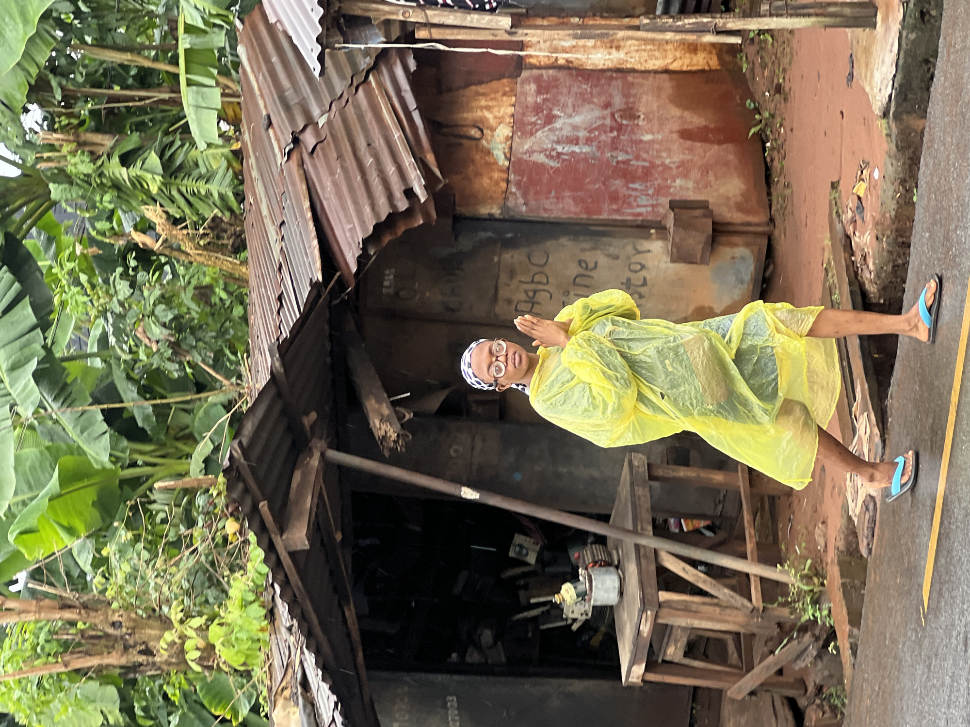 A person wearing a yellow raincoat and a headscarf stands with their hands clasped in front of them in a gesture of prayer or greeting. They are wearing glasses and flip-flops. The person is standing on a dirt path next to a corrugated metal structure with a rusted, red-painted door. The structure appears to be a stall or a small shop. To the left of the structure are large banana leaves and lush green vegetation, suggesting a tropical environment. The ground is wet and appears to have been recently rained on. The overall scene suggests an outdoor setting in a rural or semi-urban area, likely during or after rainfall. There is some visible text on the corrugated metal door, but it is difficult to read clearly. The image is oriented vertically.