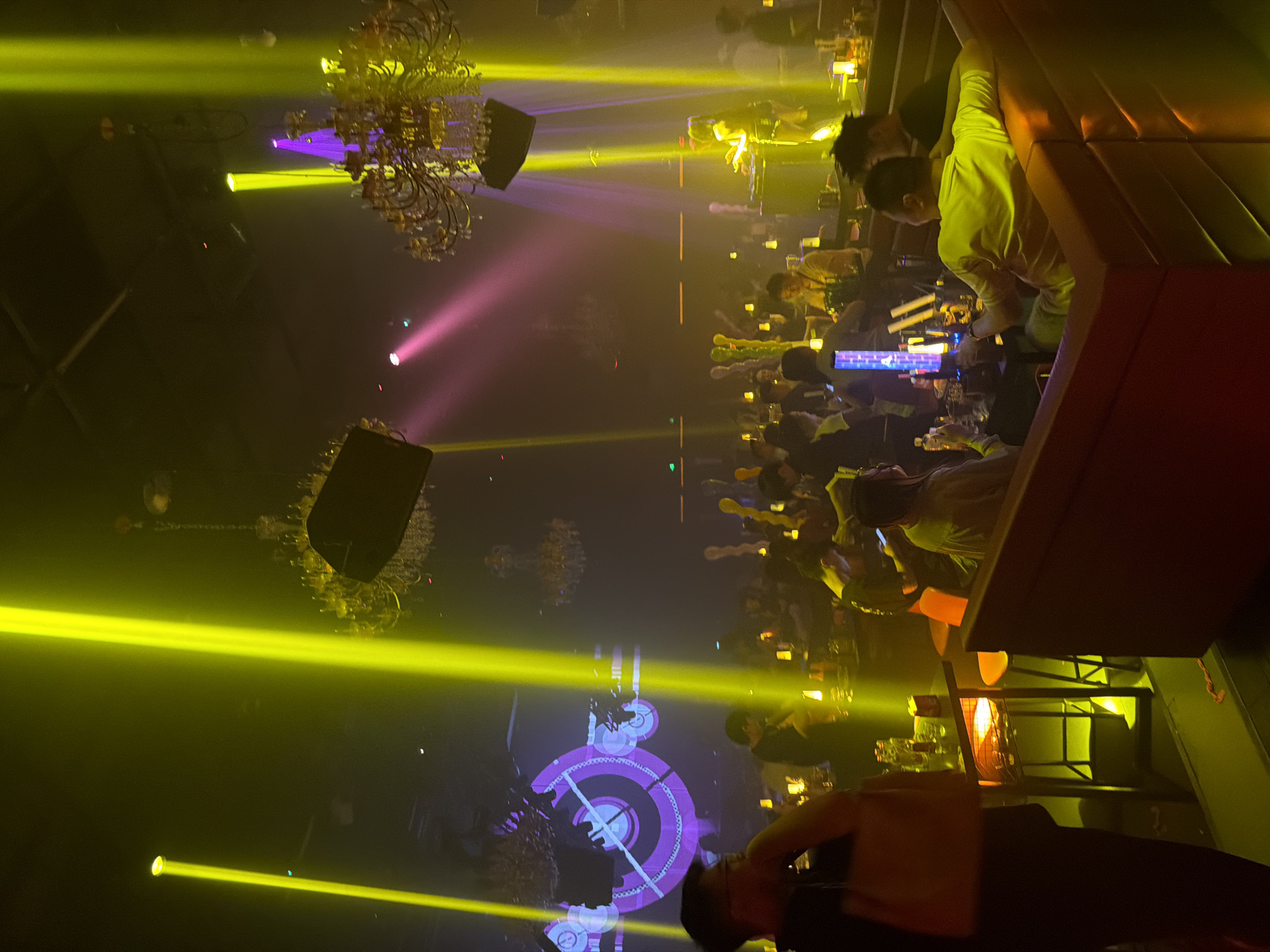 The image depicts the interior of a dimly lit nightclub with a vibrant atmosphere. Intense yellow and pink spotlights cut through the haze, illuminating a crowd of people seated at tables and interacting within the space. Elaborate chandeliers hang from the ceiling, adding to the opulent decor. In the foreground, a person in dark clothing is partially visible, holding a bag. To the right, a DJ is seen operating equipment, illuminated by the stage lighting. The scene suggests a lively social gathering with music and entertainment. The location is identified as Jiangbeicheng Street, China.