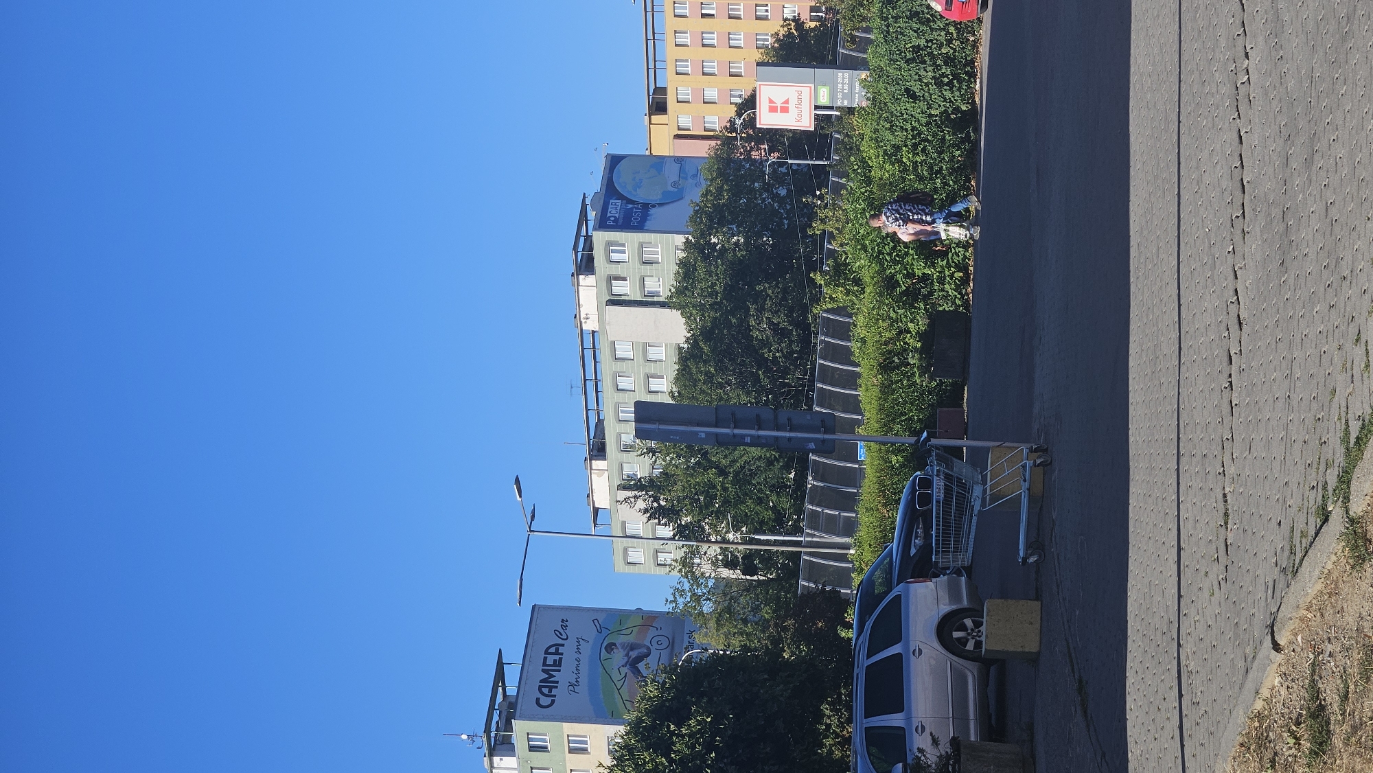 The image captures a sunny day in Prešov, Slovakia, with a clear blue sky dominating the upper portion. In the background, several multi-story apartment buildings are visible. The building on the far left has a large billboard advertisement for "CAMERA Car" with the slogan "Plníme sny". Adjacent to it is another apartment building. Further to the right, a taller, light green building stands out, and beyond that, a yellow apartment building. A red sign with the white "Kaufland" logo is positioned in front of the yellow building, indicating a supermarket.
In the foreground, a paved area resembling a parking lot or pedestrian zone is present. A silver station wagon is parked on the left side, with a black car partially visible behind it. A shopping cart is situated in the middle of the paved area, near the black car. Two individuals are walking away from the camera towards the right, with one person carrying a shopping bag. A dense hedge runs along the middle ground, separating the paved area from the buildings and trees behind. The overall scene depicts an urban environment with residential buildings, commercial signage, and everyday activity.