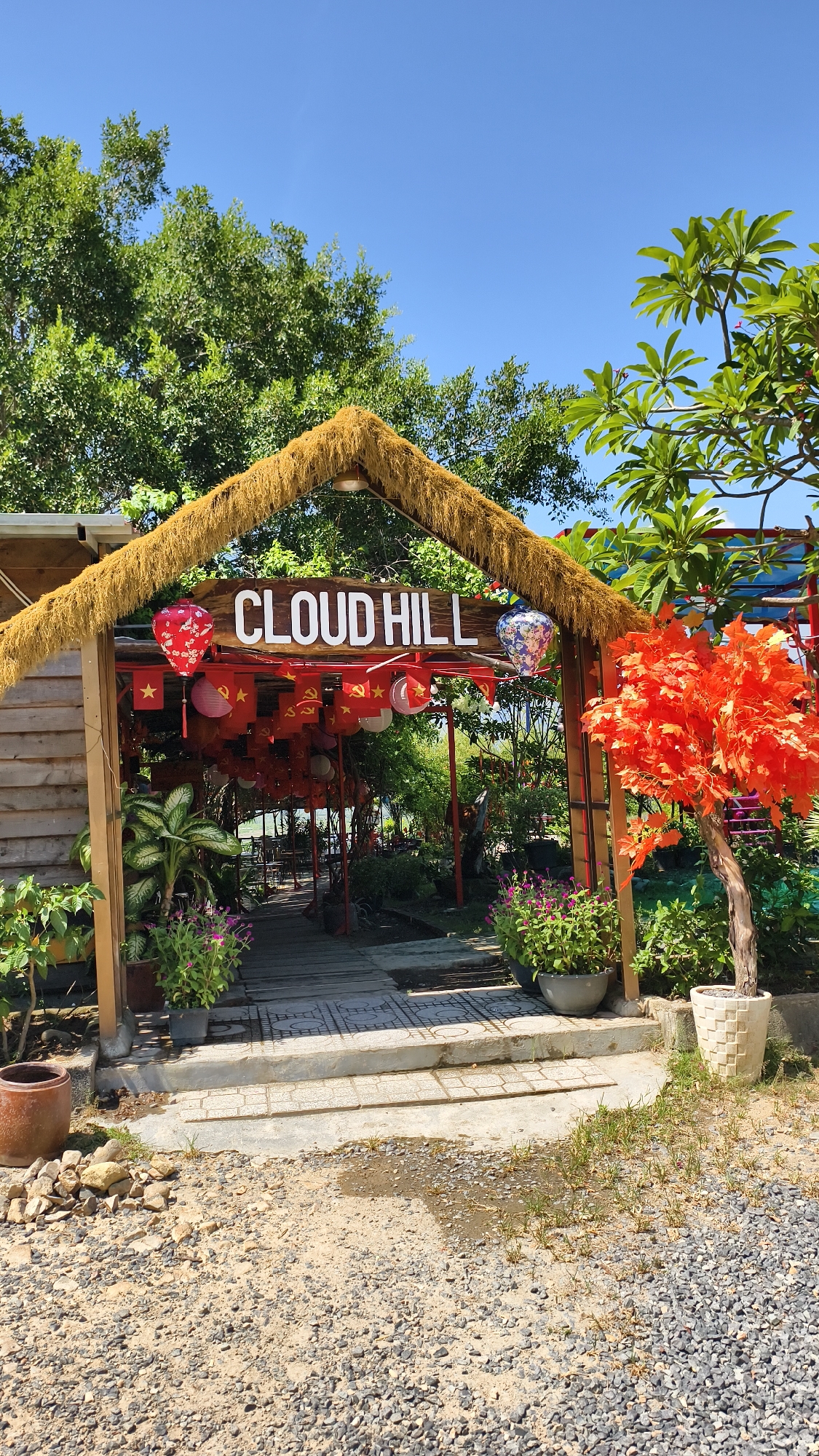 The image shows an entrance to a place called "CLOUD HILL" in Nam Nha Trang Ward, Vietnam. The entrance is a wooden structure with a thatched roof, decorated with red flags and lanterns. There are plants and flowers around the entrance. An orange artificial tree is visible to the right of the entrance. The ground in front of the entrance is covered with gravel. The sky is clear and blue. There are no people visible in the image.