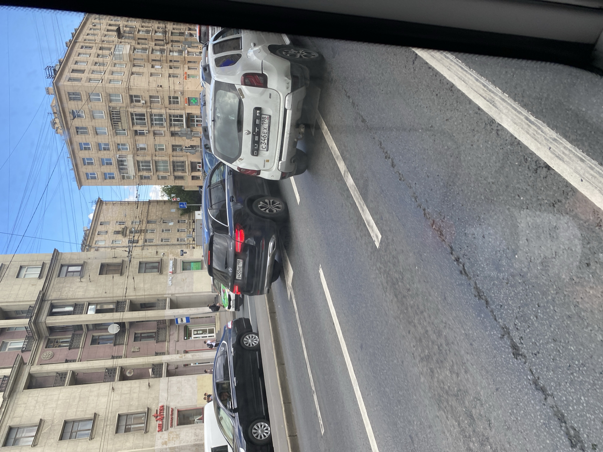 The image captures a street scene in Saint Petersburg, Russia, likely on a sunny day with some cloud cover. In the foreground, the asphalt road shows clear lane markings, including white dashed lines, and some areas of repair or wear. On the right side, a white Renault Duster SUV with a license plate starting with "C346EX" is visible, facing away from the camera. To its left, a dark Mercedes-Benz SUV with a license plate starting with "B450HE" is also seen. Further to the left, a black sedan is observed in traffic.
The background features multi-story buildings with characteristic St. Petersburg architecture, including numerous windows and balconies. Some of these buildings have commercial establishments on the ground floor, with signage in Russian, such as "MILAVITSA" and a green cross indicating a pharmacy. Power lines and streetlights are also present, indicating an urban environment. A few pedestrians are visible on the sidewalk, and a green and white vehicle, possibly a taxi or public transport, is further down the street. The overall impression is that of a typical urban street with moderate traffic.
