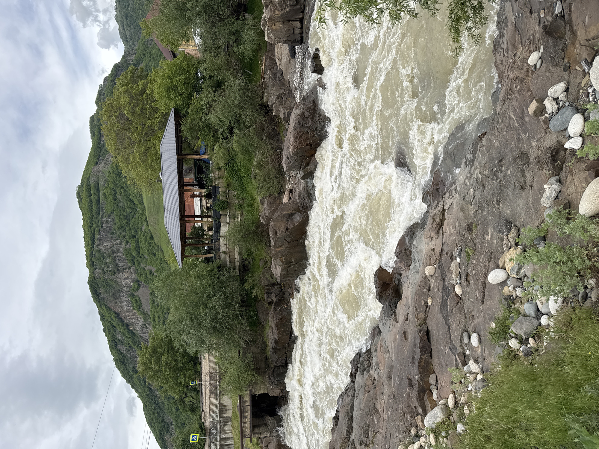 A river flows through a rocky landscape in Каменномостский, Russia. The river, a turbulent, opaque white, dominates the right half of the frame. The riverbanks are a mix of dark brown rocks of various sizes and light-colored pebbles. Small patches of green vegetation grow along the edges of the rocks.
To the left of the river is a vegetated hill. A structure with a grey roof, likely a gazebo or covered seating area, is situated on the hill. At the base of the hill is a concrete wall, which abuts a road with a pedestrian crossing sign. The sky is overcast.