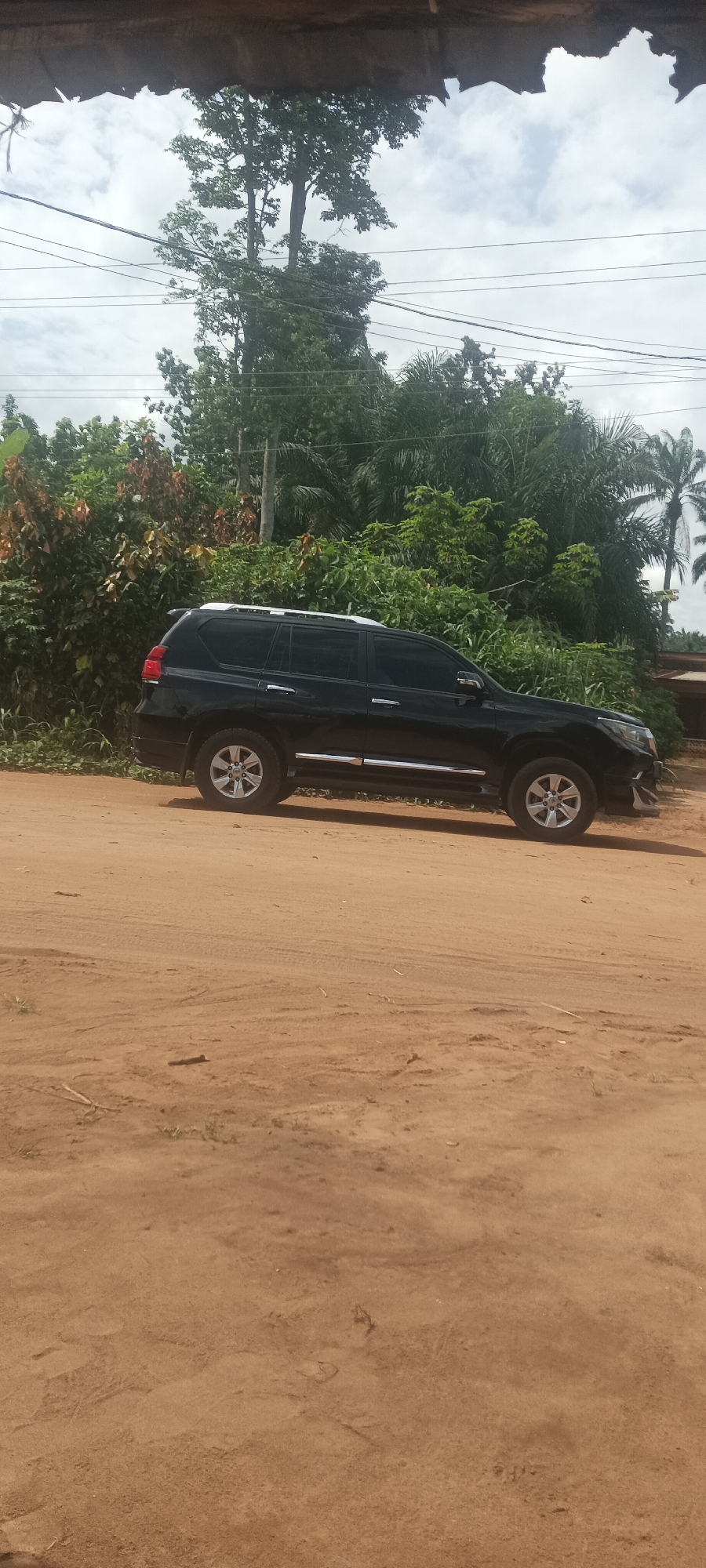 A black Toyota Land Cruiser Prado is parked on a reddish-brown dirt road in Amauzu, Nigeria. The vehicle is positioned in profile, facing right. The surrounding environment features dense vegetation, including trees and shrubs. Overhead, utility lines are visible against a cloudy sky. A partially visible roof structure overhangs the top of the frame.